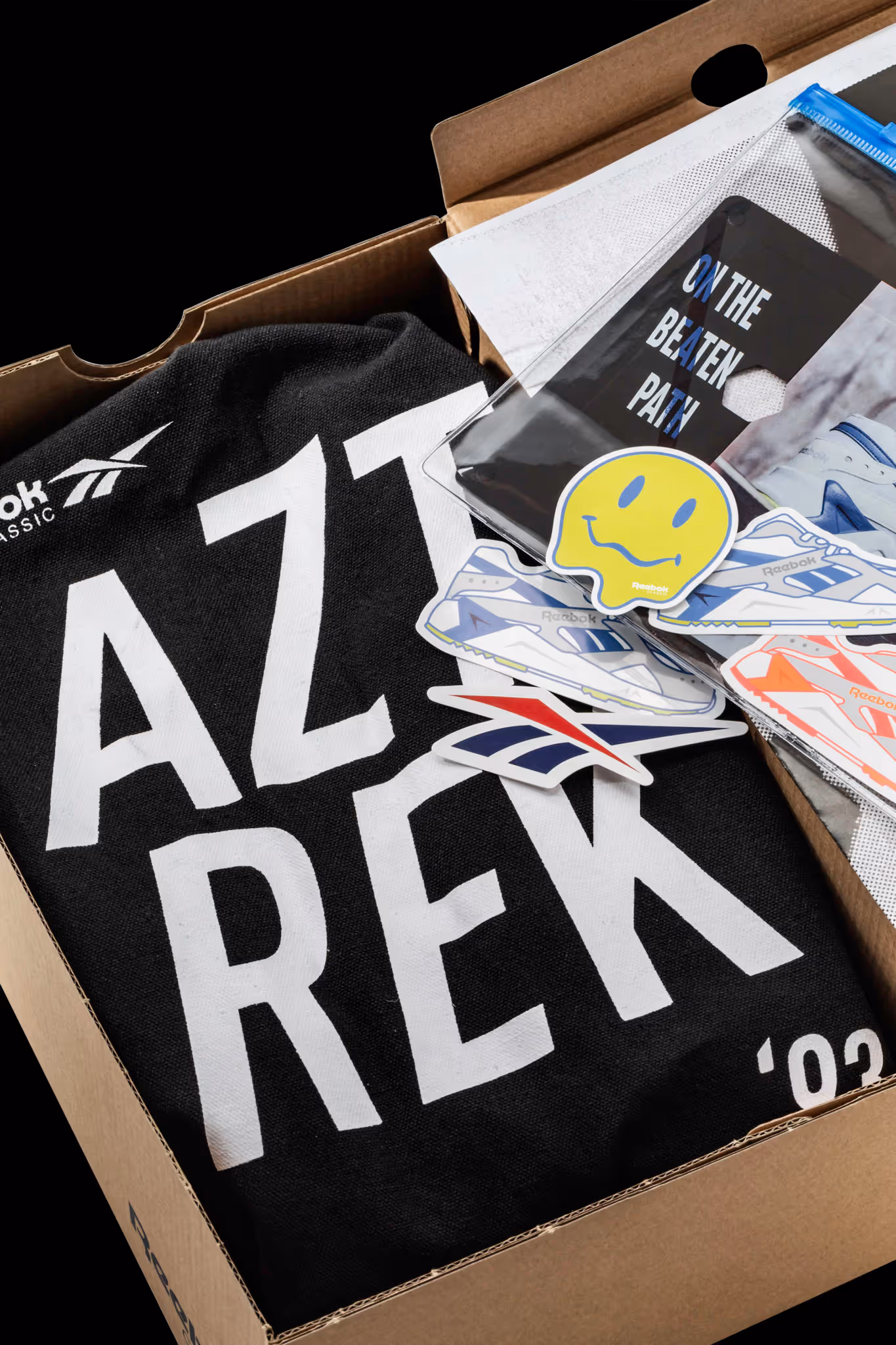 Open cardboard box containing a black Reebok Classic shirt with 'AZT REK' text and various Reebok-themed stickers including a yellow smiley face and sneaker designs.