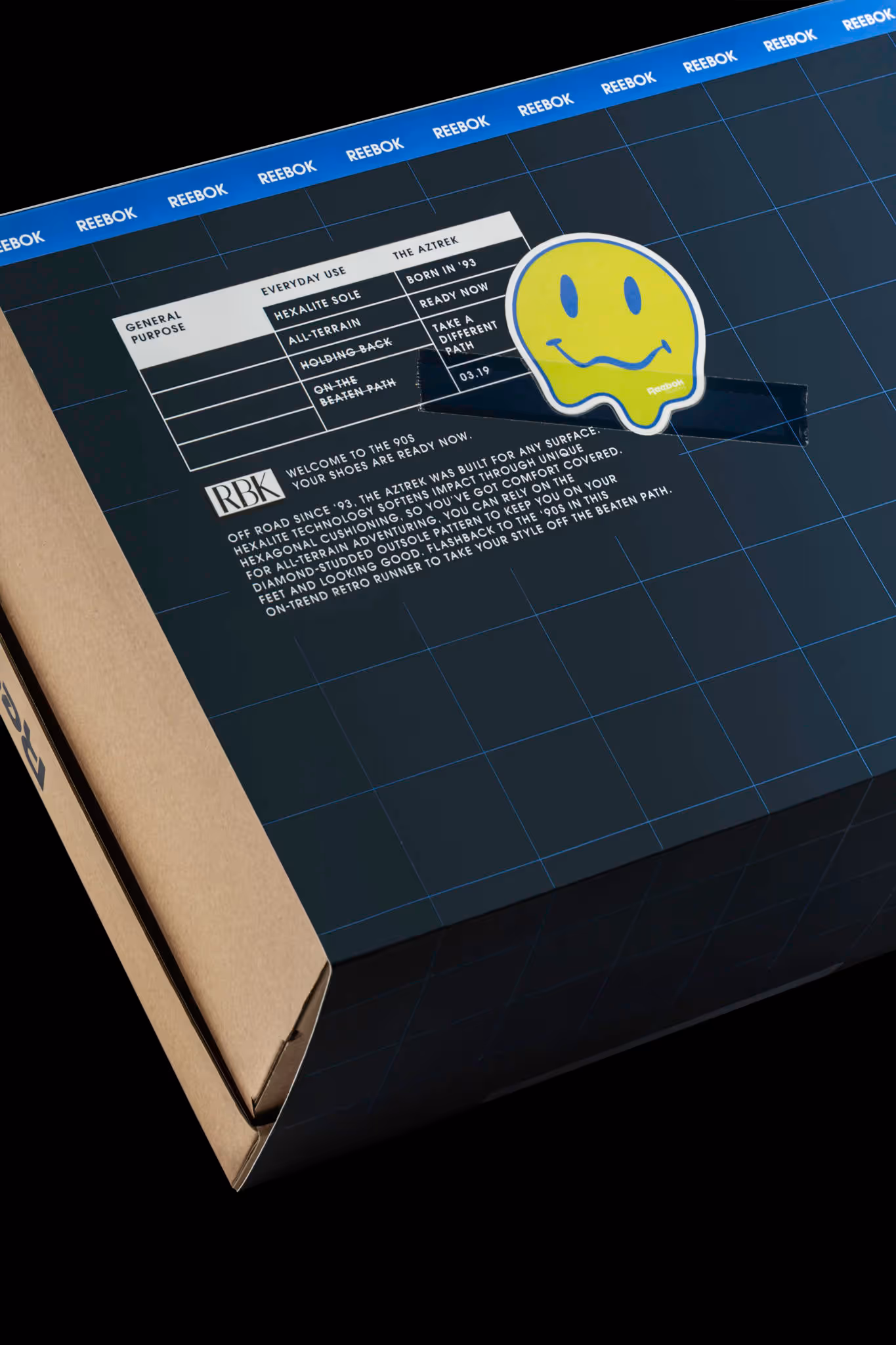Reebok shoe box with a blue grid design, product information chart, and a yellow smiley face sticker.
