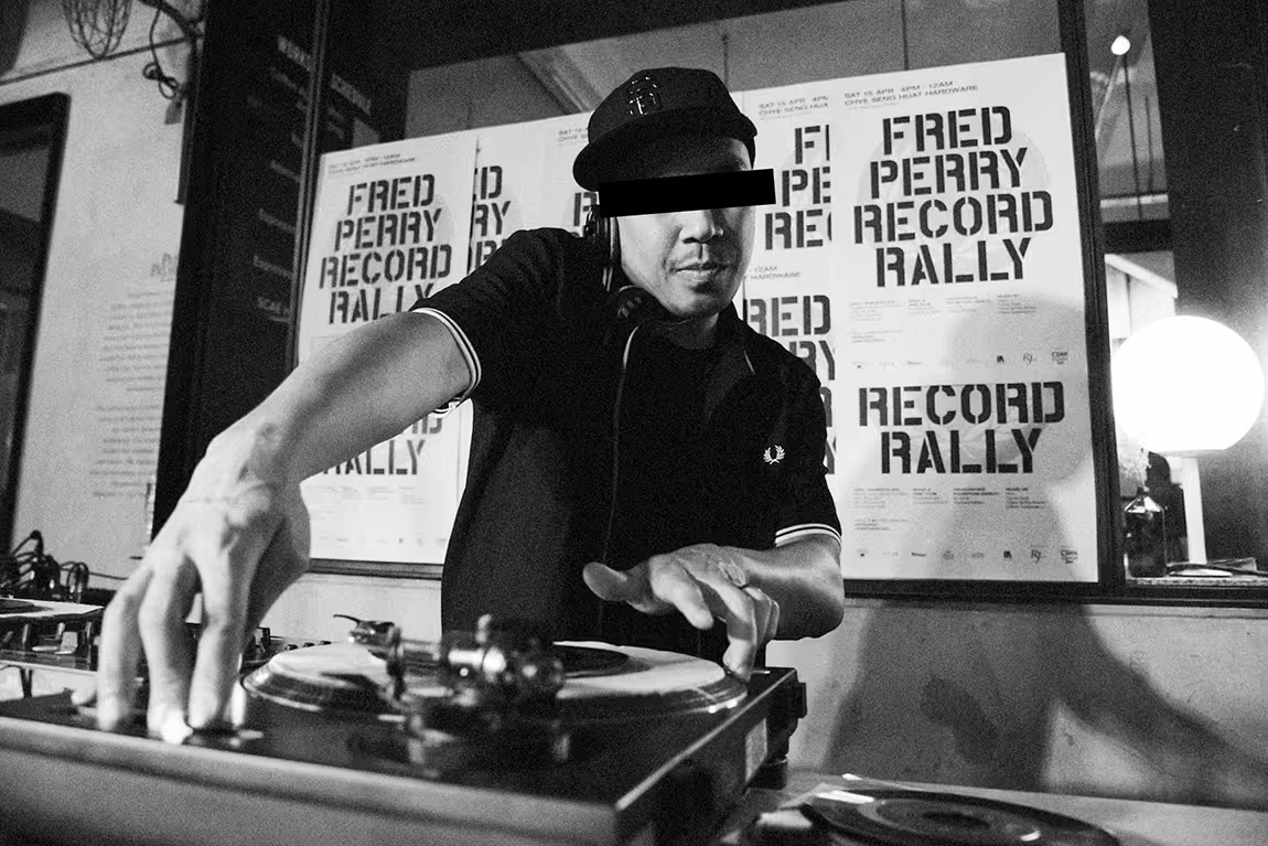 Black and white photo of a DJ wearing a cap, spinning vinyl records with 'Fred Perry Record Rally' posters in the background.