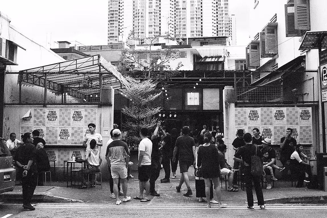 Black and white photo of a street scene with a group of people gathered outside a building with posters reading 'Fred Perry Record Rally'.