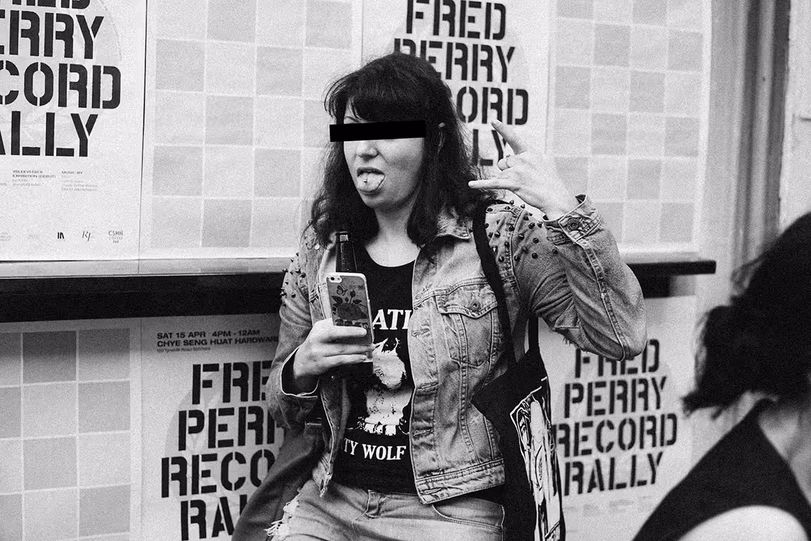 Person with black bar over eyes sticking out tongue and making rock hand gesture, wearing studded denim jacket and holding a phone in front of posters that say Fred Perry Record Rally.