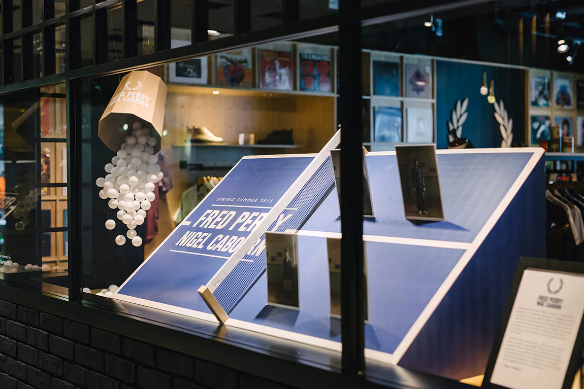 Retail display window featuring a tilted blue ping pong table branded with Fred Perry and Nigel Cabourn, white ping pong balls spilling from a branded container above, and framed photos on the table.