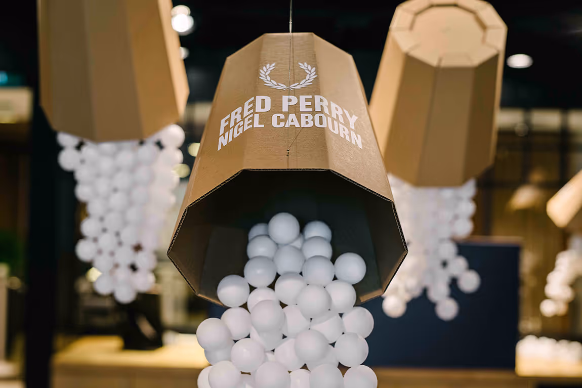 Hanging cardboard containers labeled Fred Perry Nigel Cabourn spilling clusters of white balls in an indoor setting.