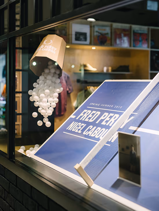 Store window display with a tilted cup pouring white ping pong balls onto a blue ping pong table featuring Fred Perry and Nigel Cabourn branding.