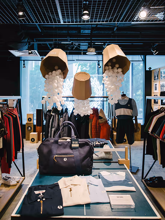 Clothing store display featuring folded polo shirts, a black duffel bag, and hanging outerwear, with decorative cardboard containers pouring out white balls overhead.