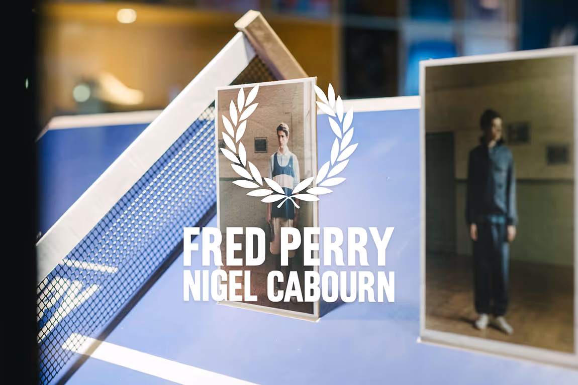 Fred Perry Nigel Cabourn logo overlaid on blurred photos of men wearing casual clothing in an indoor setting.