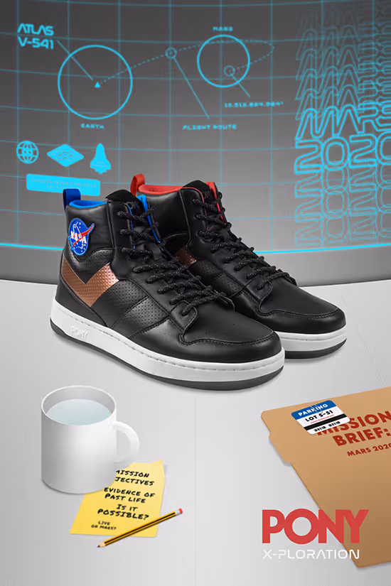 Pair of black high-top sneakers with NASA patch and copper chevron detail, displayed on a surface with a white mug, yellow notes, pencil, and a brown folder labeled Mission Brief Mars 2020.