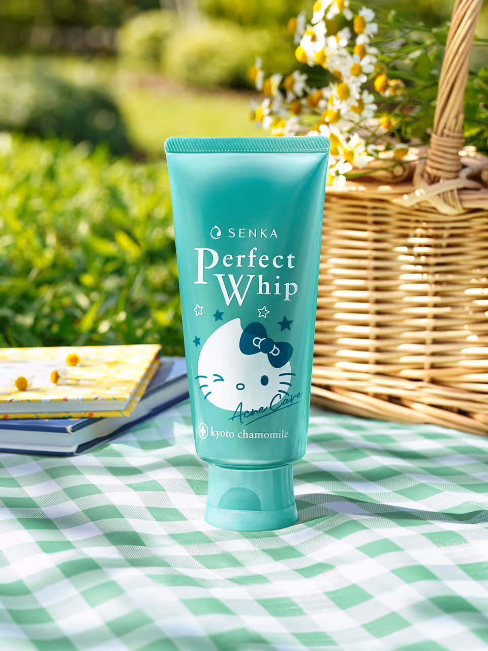 Tube of Senka Perfect Whip Acne Care facial cleanser with Hello Kitty design on a green and white checkered cloth, beside a wicker basket and notebooks outdoors.