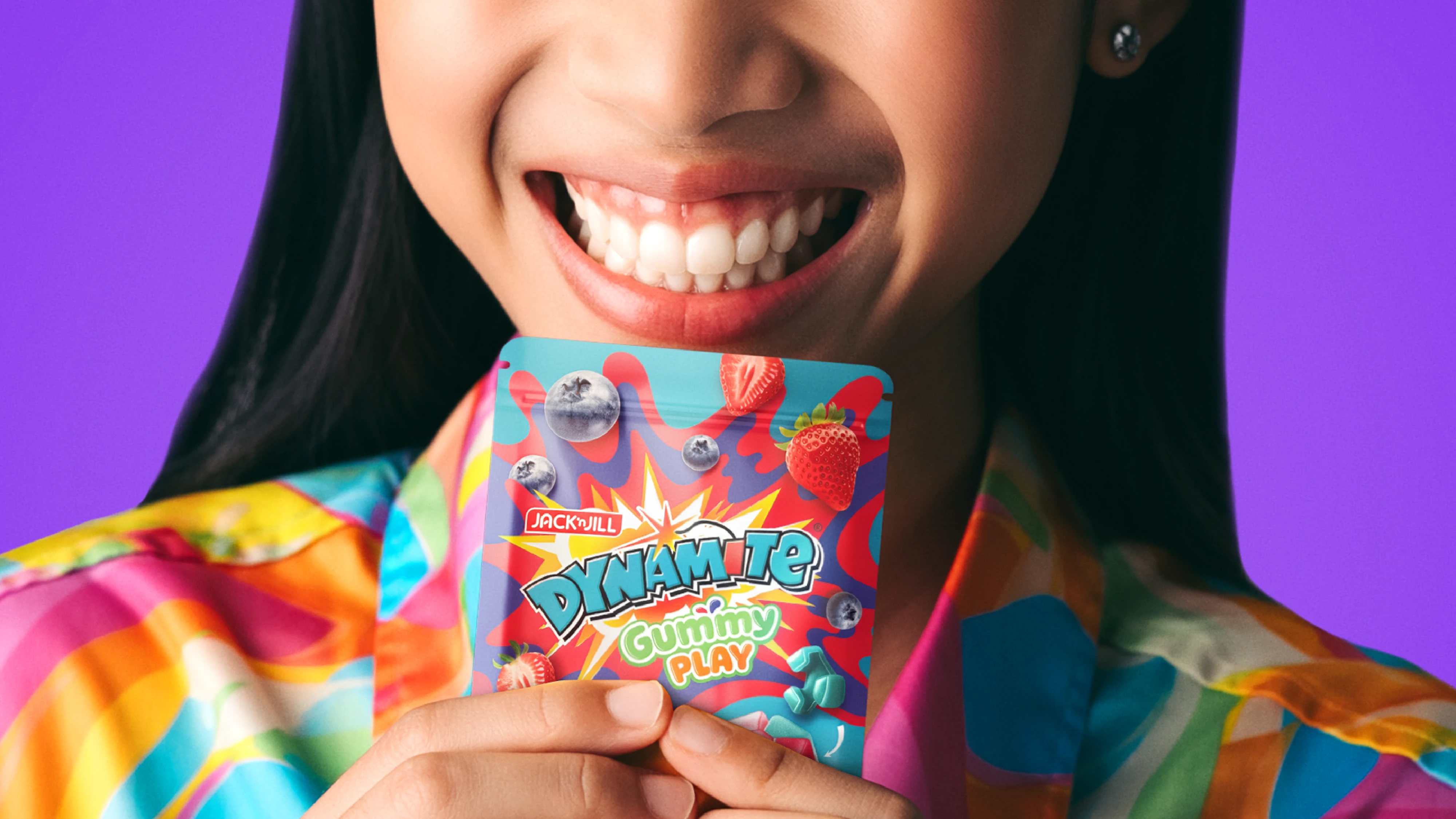 Smiling person holding a colorful package of Jack 'n Jill Dynamite Gummy Play candy.