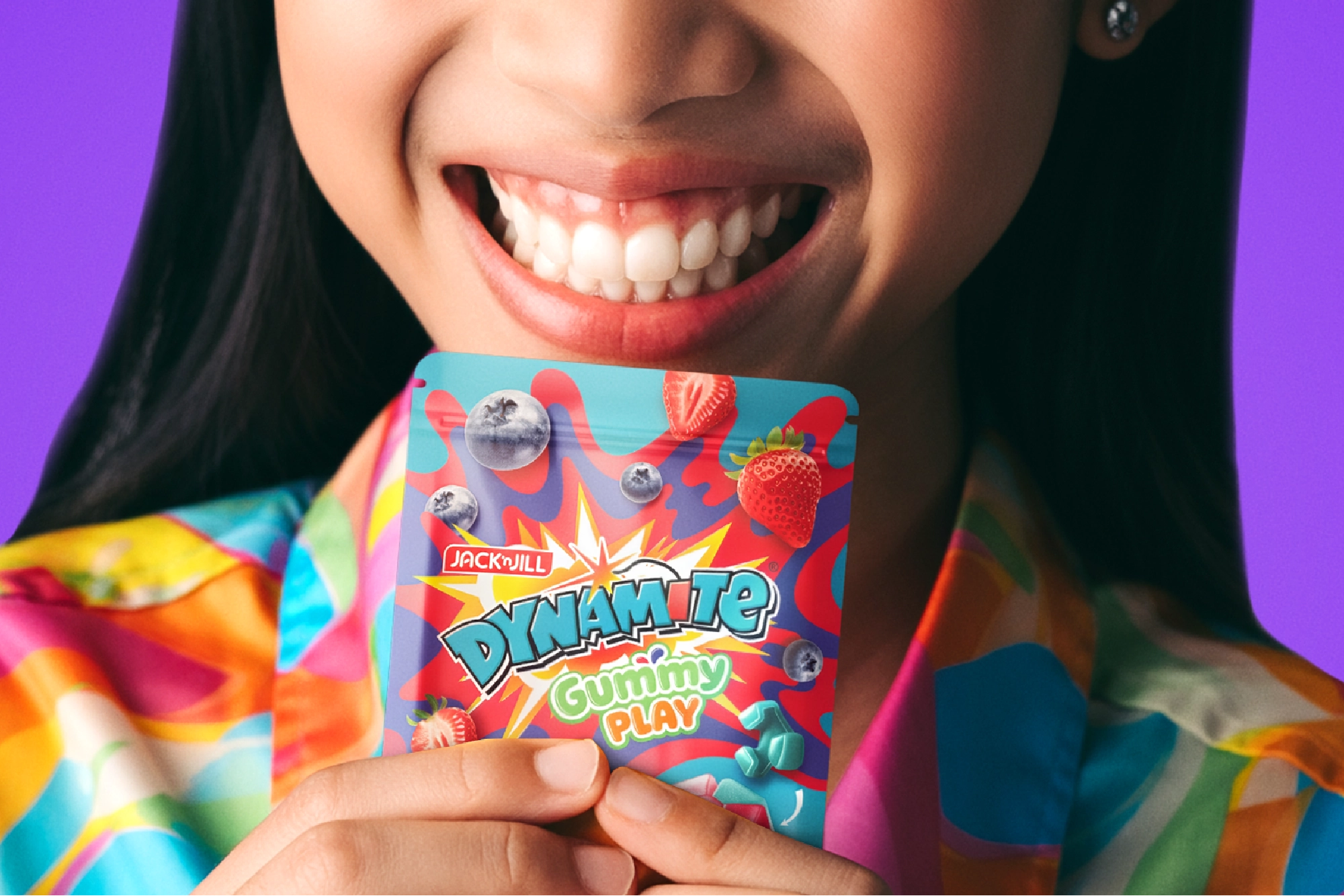 Close-up of a smiling person holding a colorful packet of Jack'n Jill Dynamite Gummy Play candy with blueberries and strawberries on the packaging.