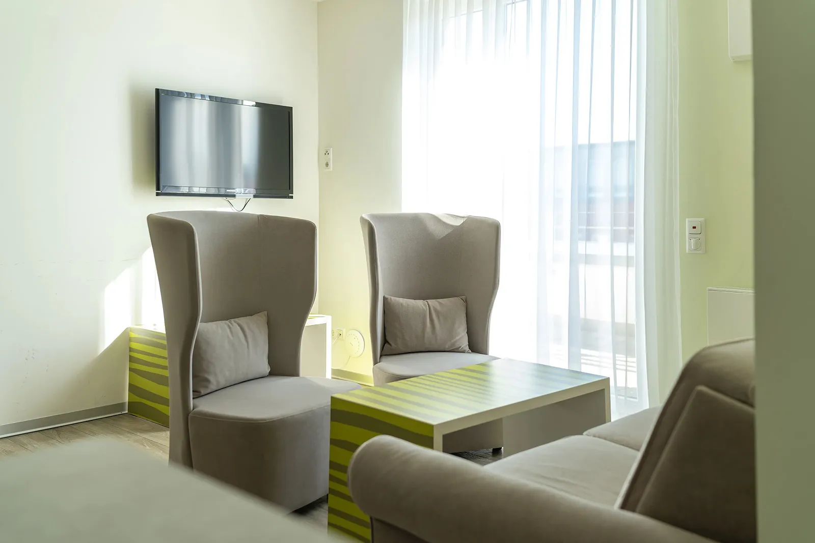 Bright seating area with two beige wingback chairs, a green-striped coffee table, beige sofa, and wall-mounted TV.
