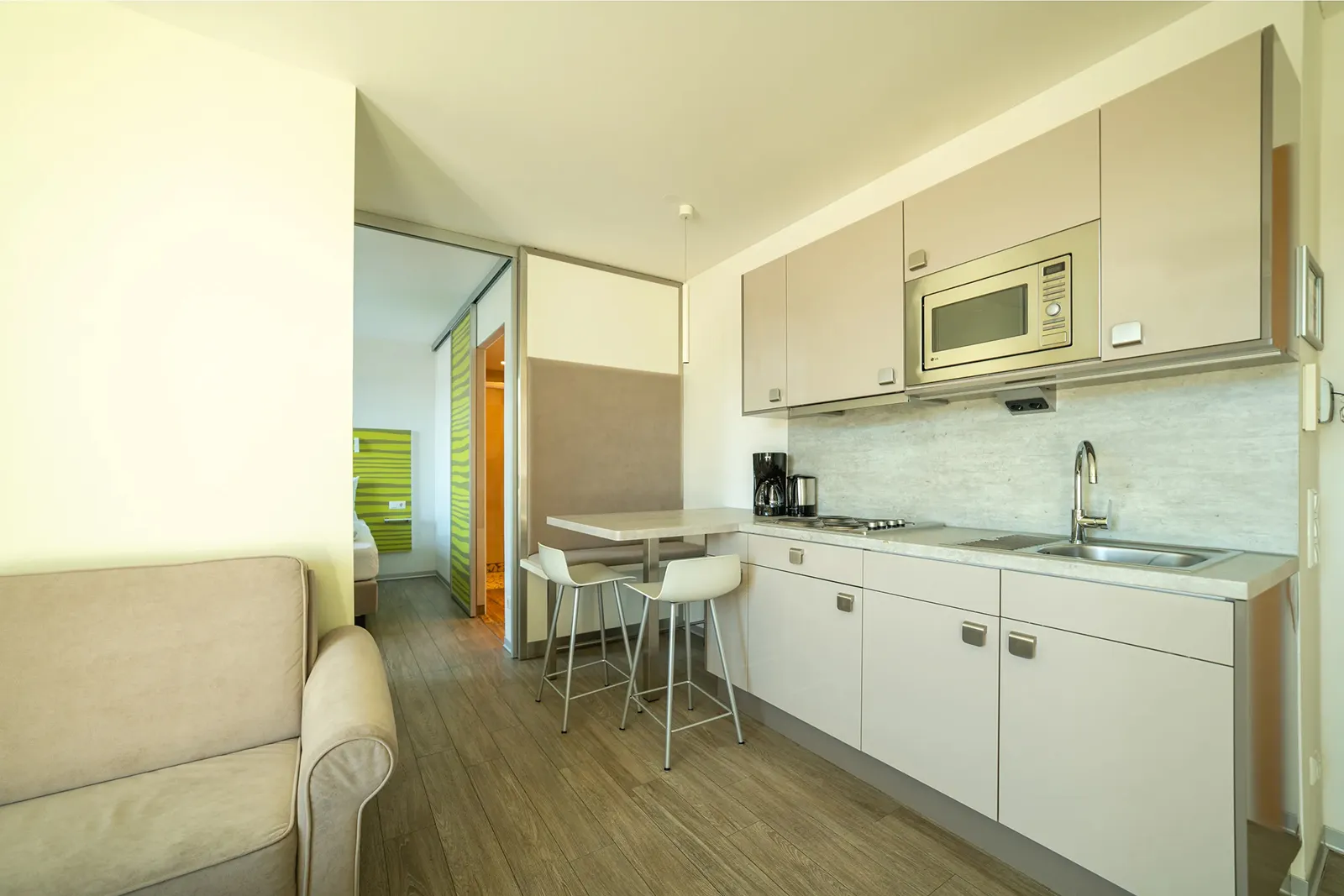 Modern kitchenette with white cabinets, built-in microwave, sink, stovetop, coffee maker, small dining table with two white bar stools, and a beige sofa visible on the left.