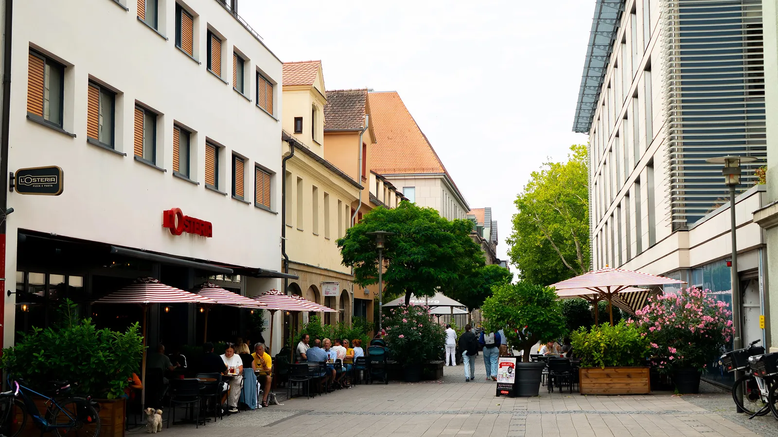 Outdoor dining area of L'Osteria restaurant with people seated under striped umbrellas along a pedestrian street lined with greenery and buildings.