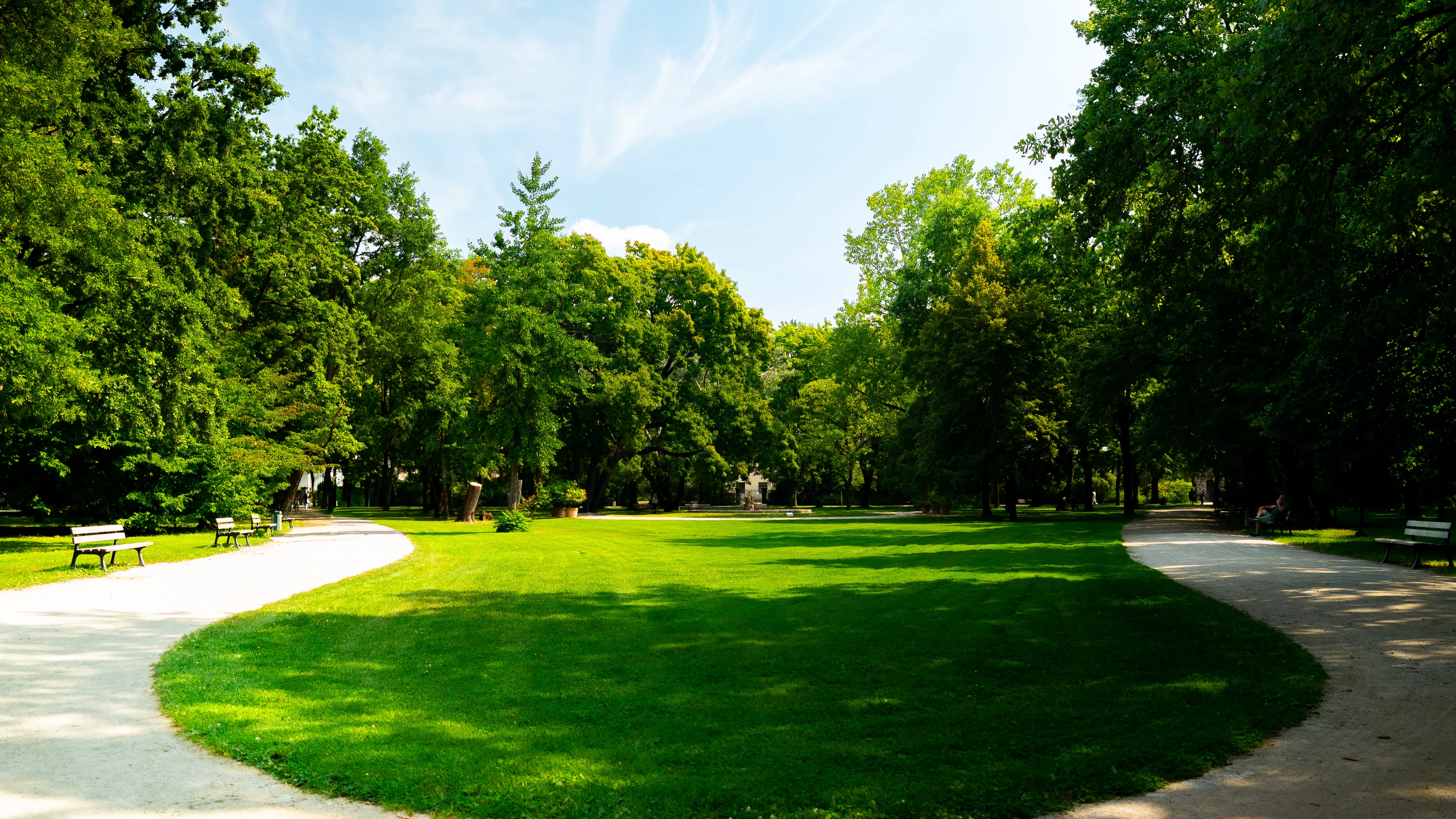 Wide green park lawn with curved walking paths and benches under a partly cloudy sky.