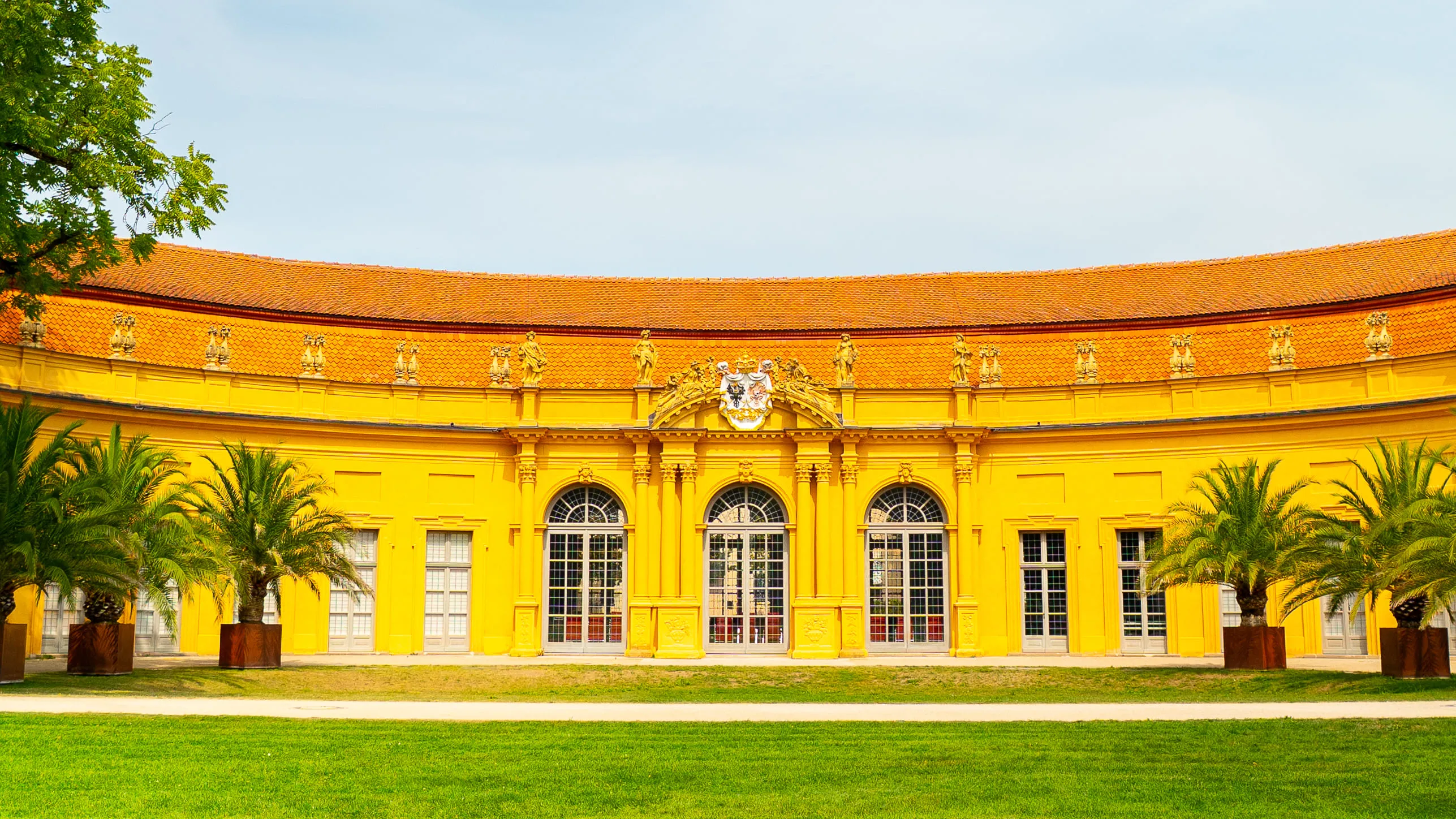 Yellow curved building with large arched windows, orange tiled roof, ornamental details, and palm trees in front.
