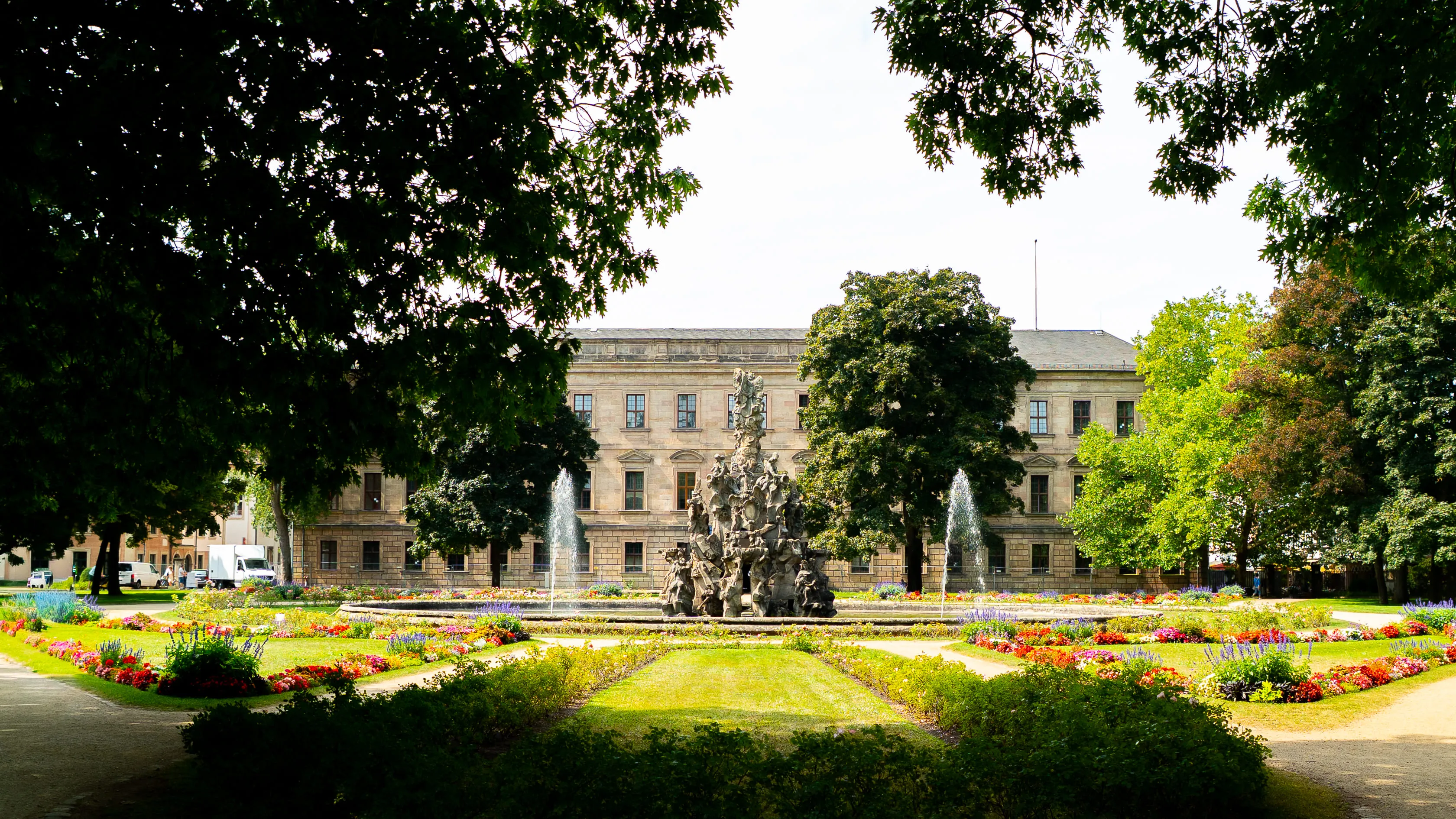 Ornate stone fountain with statues in a formal garden with colorful flower beds and large trees, set in front of a historic building.