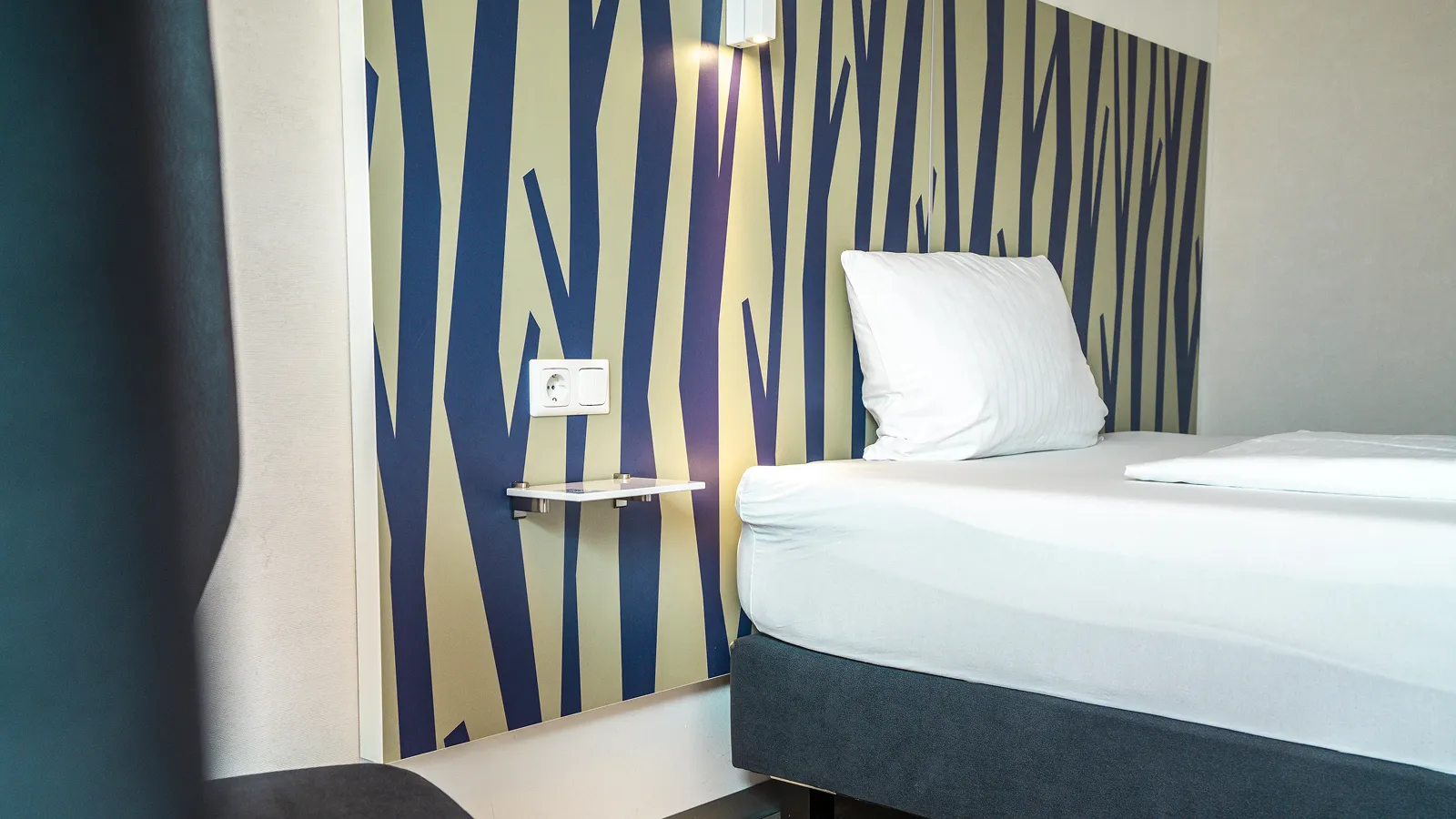 Modern hotel room corner with a bed, white pillow, wall-mounted reading light, power outlet, and patterned headboard.