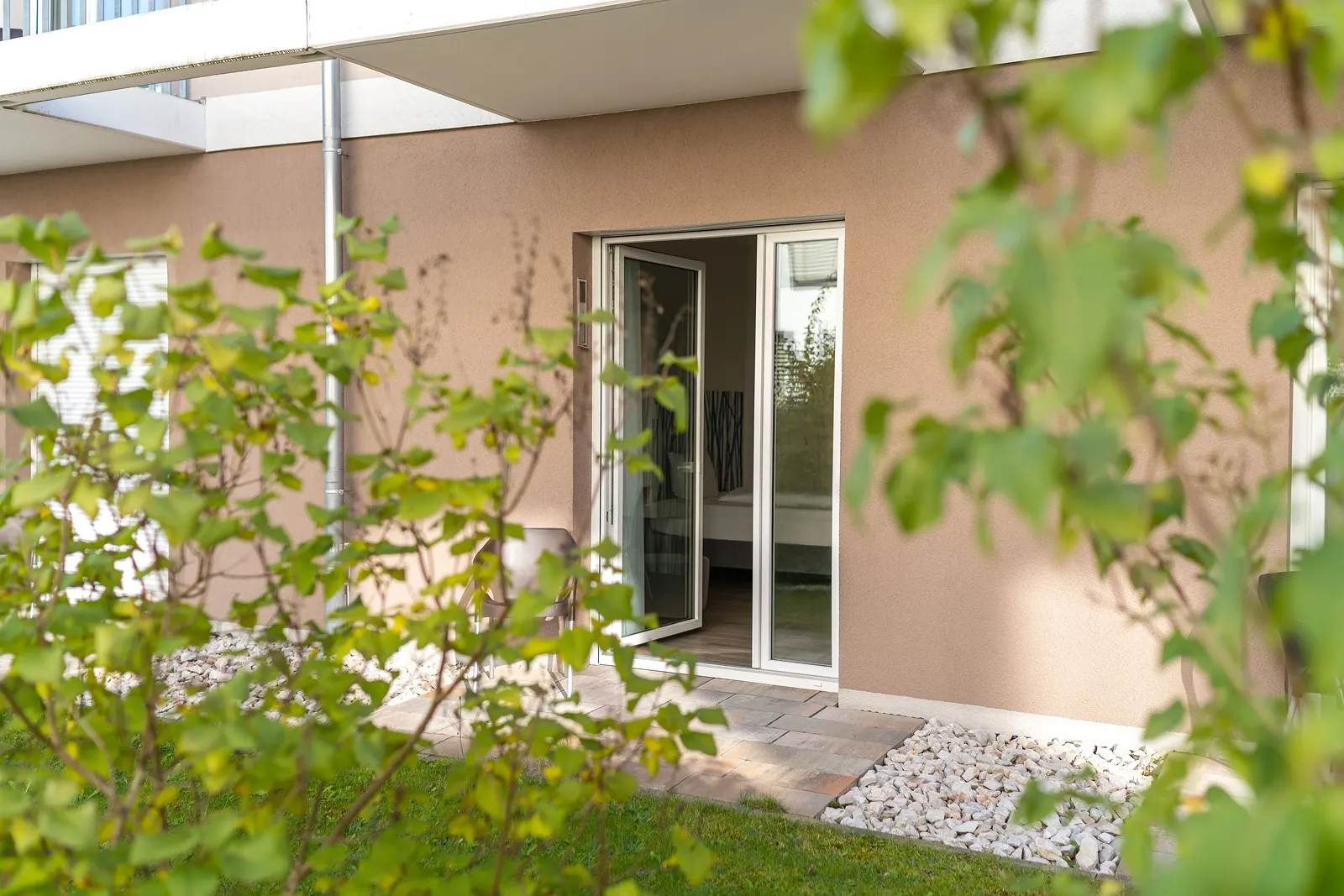 Open glass door leading to a modern bedroom patio with greenery and stone landscaping outside.