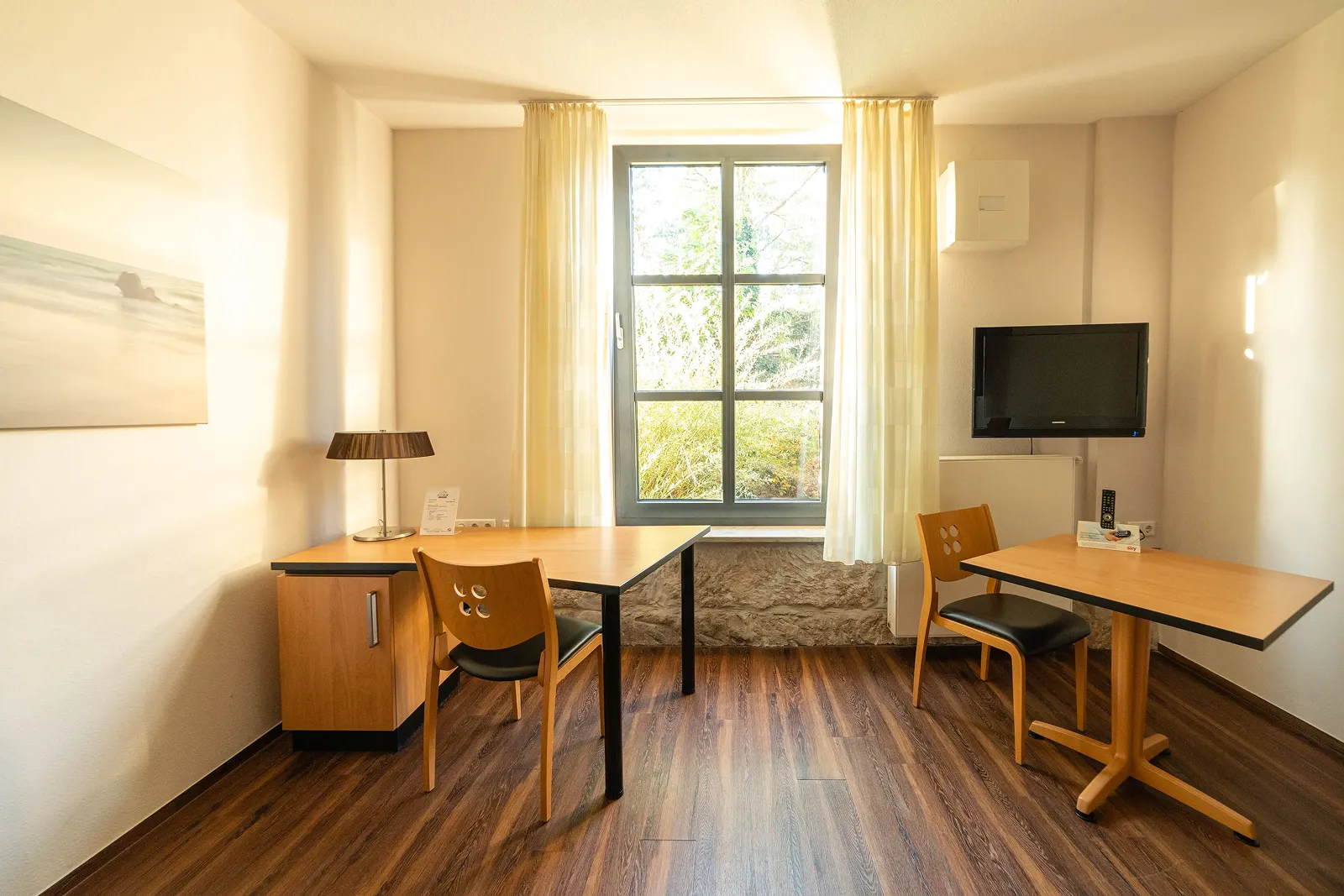 Bright room with two wooden tables, two chairs, a window with light curtains, a wall-mounted TV, and a table lamp on the left table.