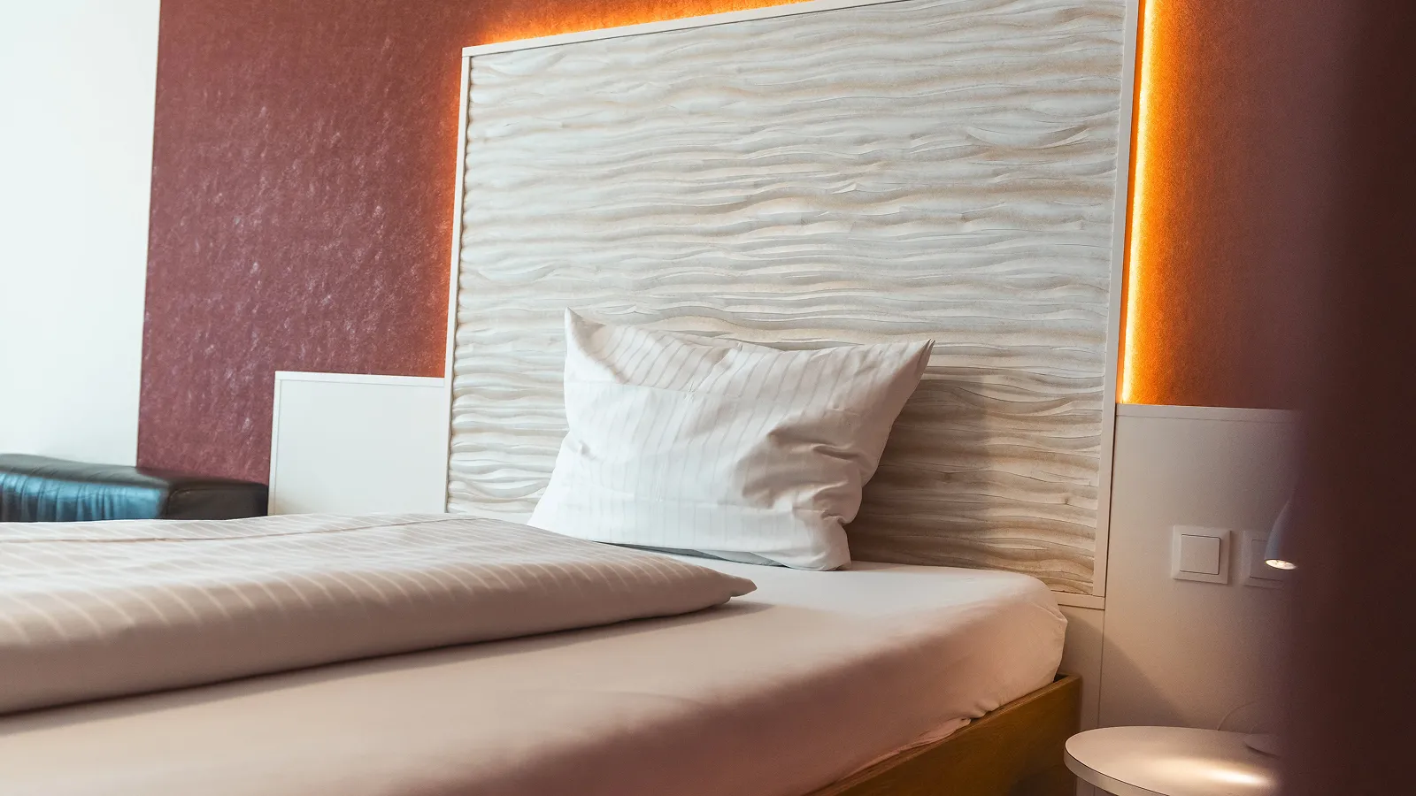 Close-up of a neatly made bed with a textured wooden headboard, white striped pillow and duvet in a modern room.