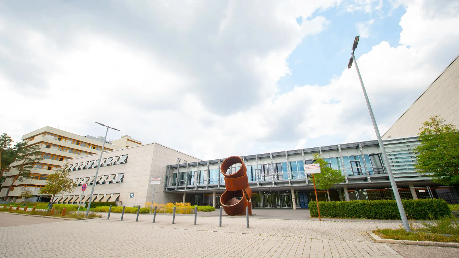 Modern building with large windows and a rust-colored abstract sculpture in front, under a cloudy sky.