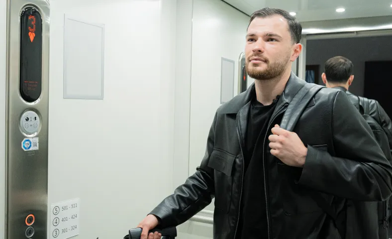 Man in black leather jacket with backpack standing inside an elevator near the control panel.