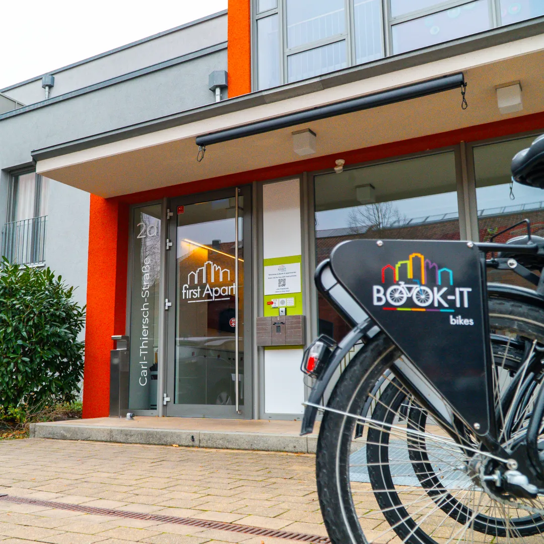 Entrance to a modern apartment building with glass doors labeled 'first Apart' and shared BOOK-IT bike parked outside.