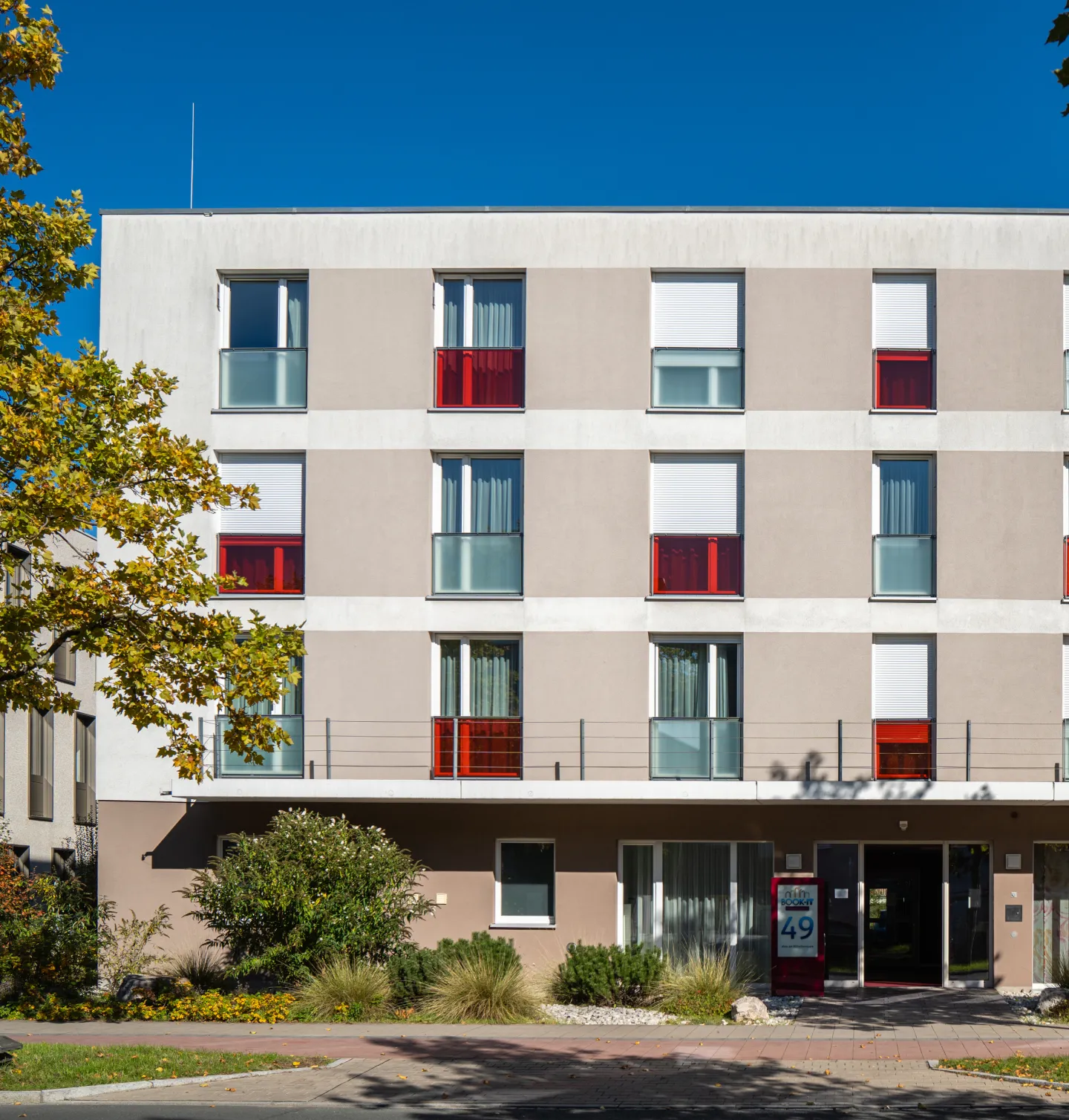 Modern three-story building with beige and white facade featuring windows with red and frosted glass panels and greenery in front.