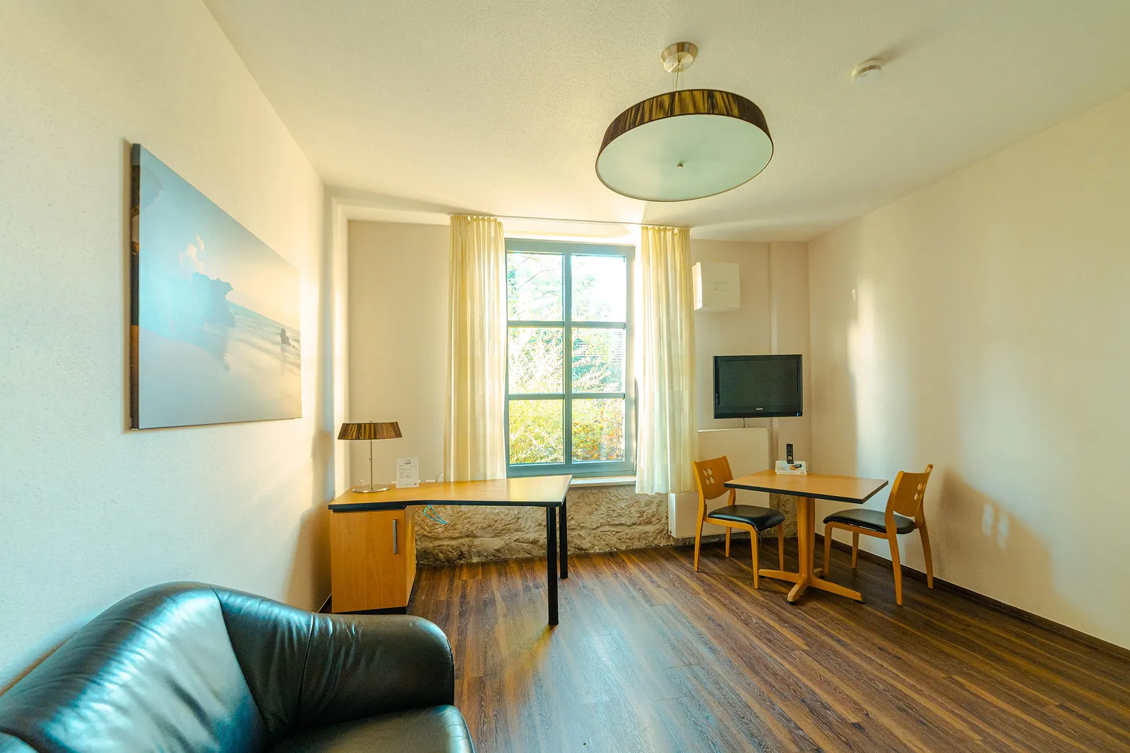 Bright room with a large window, wooden floor, black leather armchair, two wooden chairs at a small table, a desk with a lamp, and a wall-mounted TV.