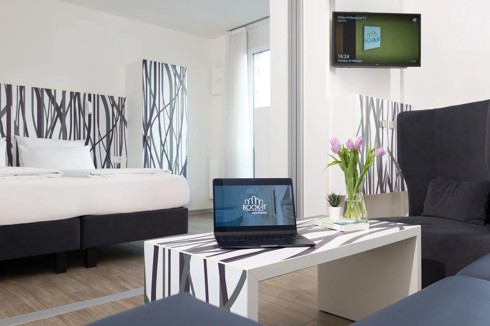Modern hotel room with a bed, patterned furniture, a laptop on the table displaying BOOK-IT apartments logo, a vase of pink tulips, and a wall-mounted TV.