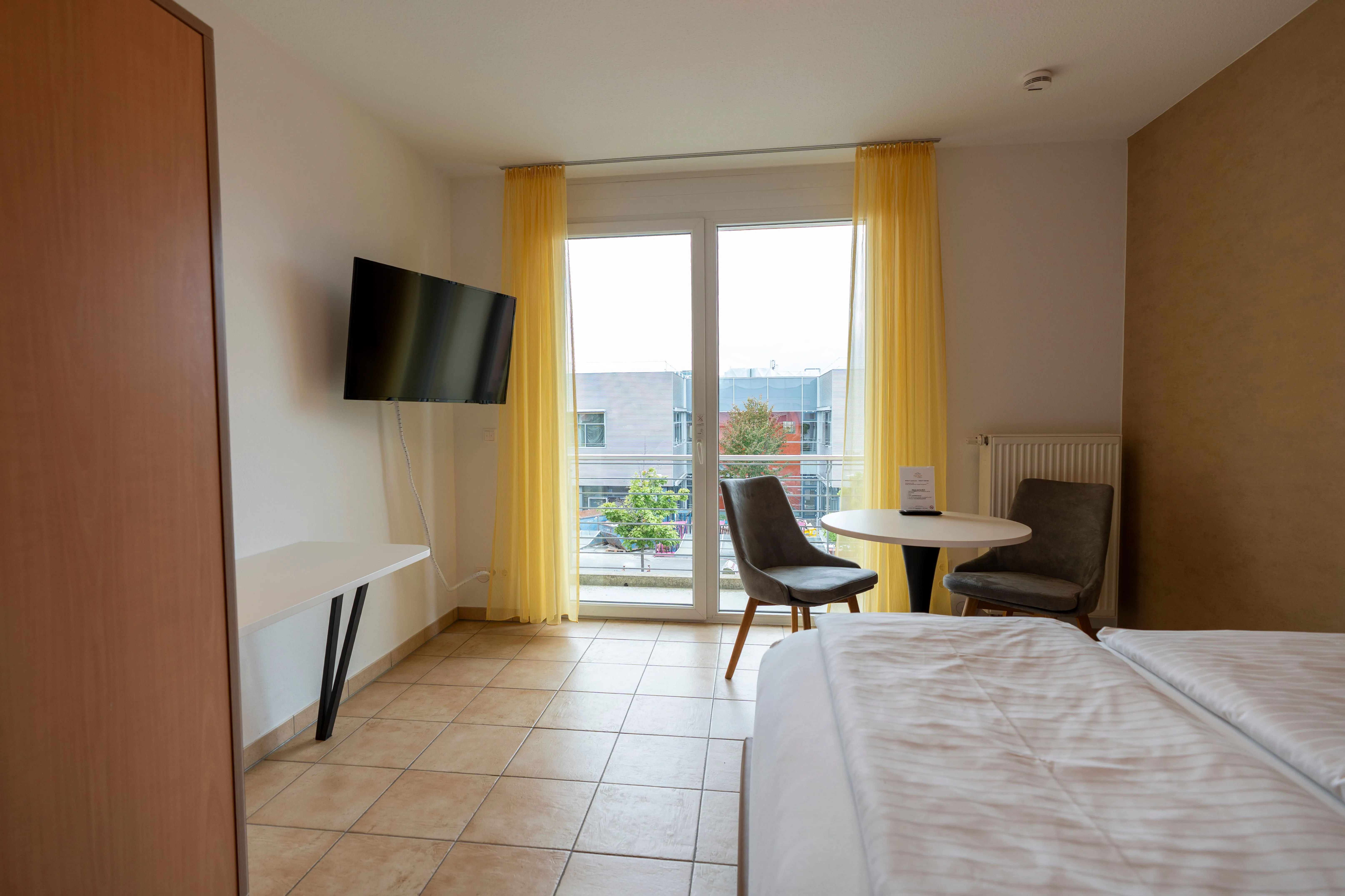 Hotel room with two single beds, a wall-mounted TV, a round table with two gray chairs near a glass door with yellow curtains, and tiled floor.