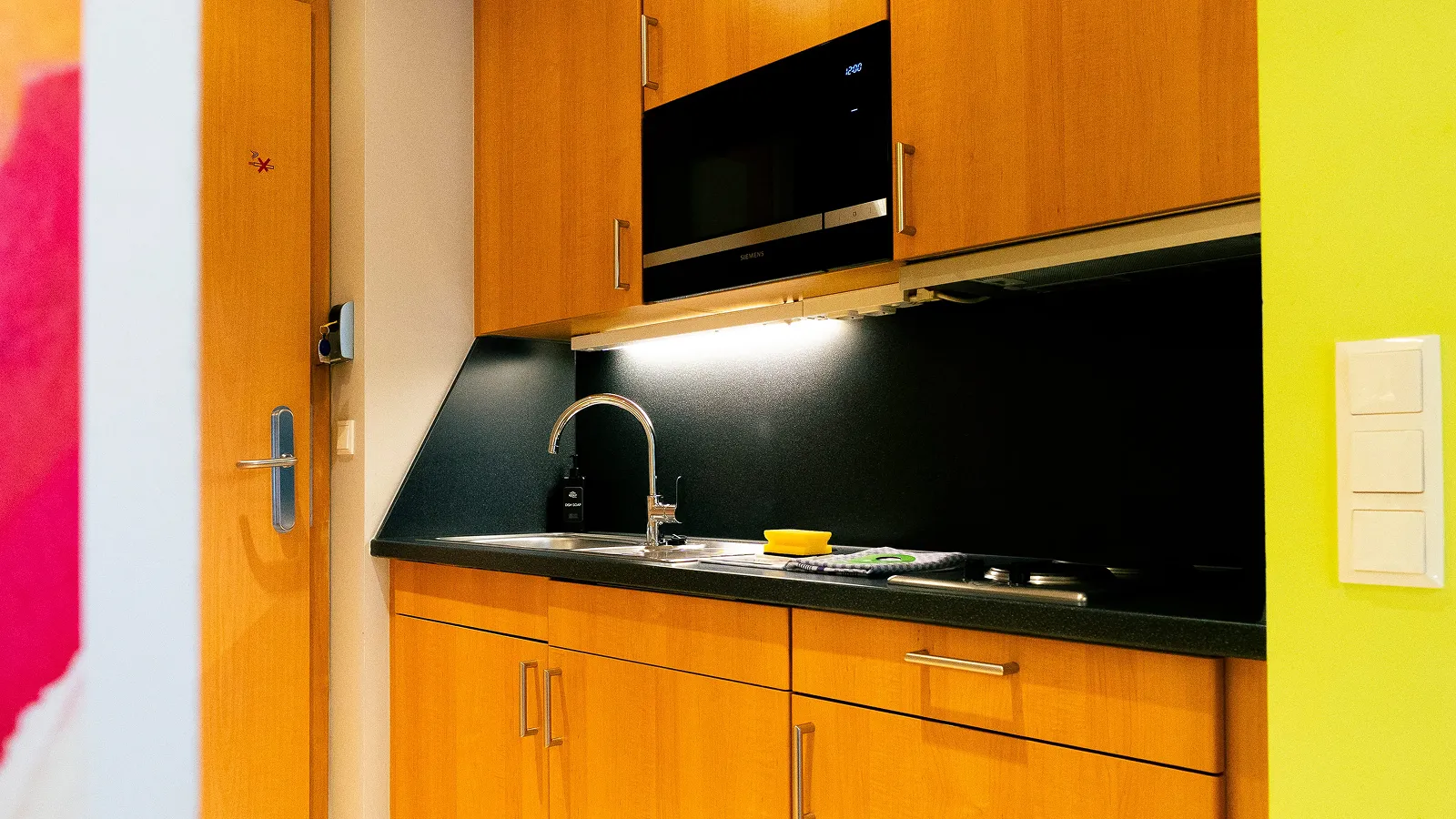 Compact kitchen area with wooden cabinets, a stainless steel sink with a faucet, a black countertop, a microwave, and cleaning items including a sponge and dish soap.