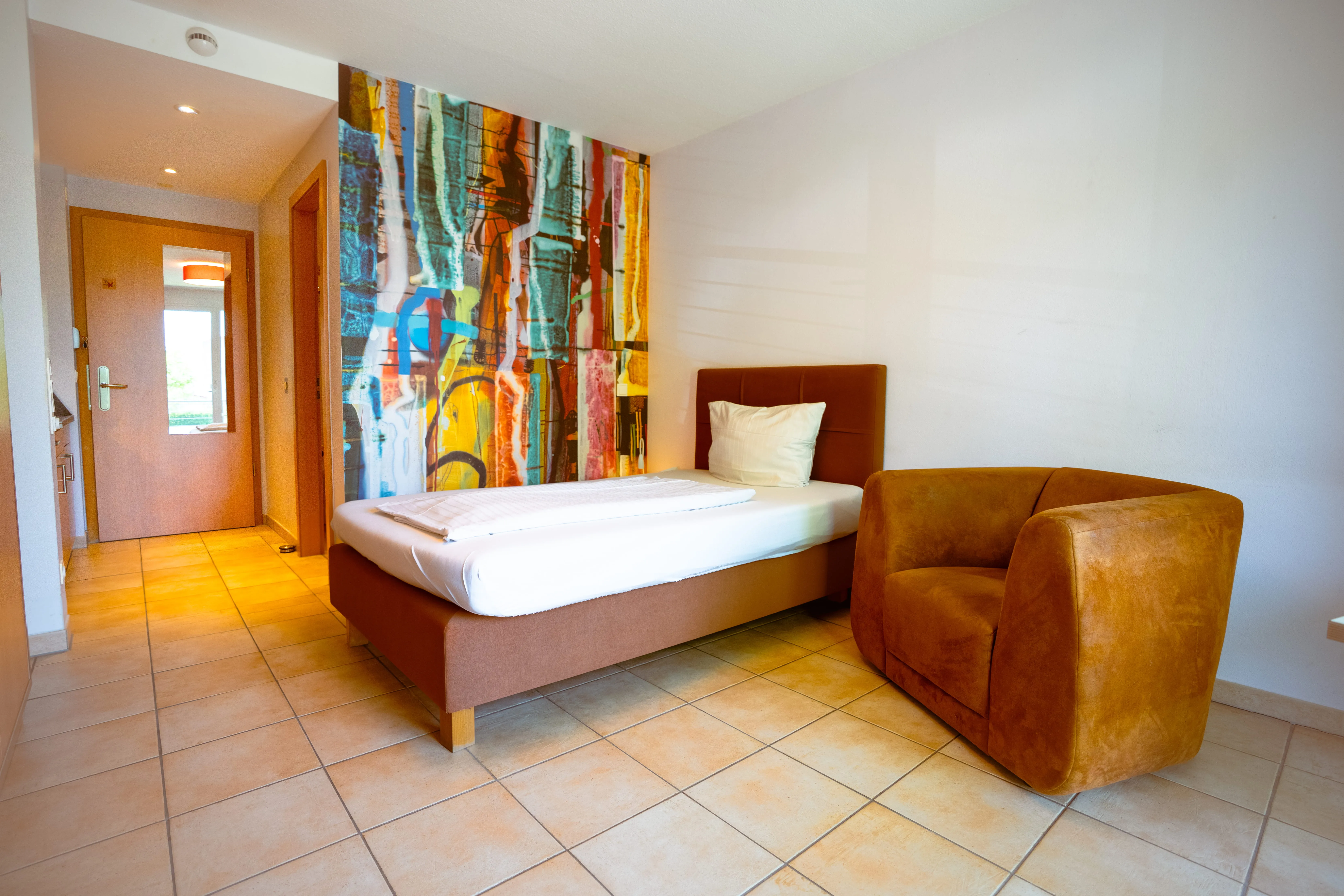 Simple bedroom with a single brown bed, a colorful abstract mural behind, and a mustard yellow armchair on tiled floor.