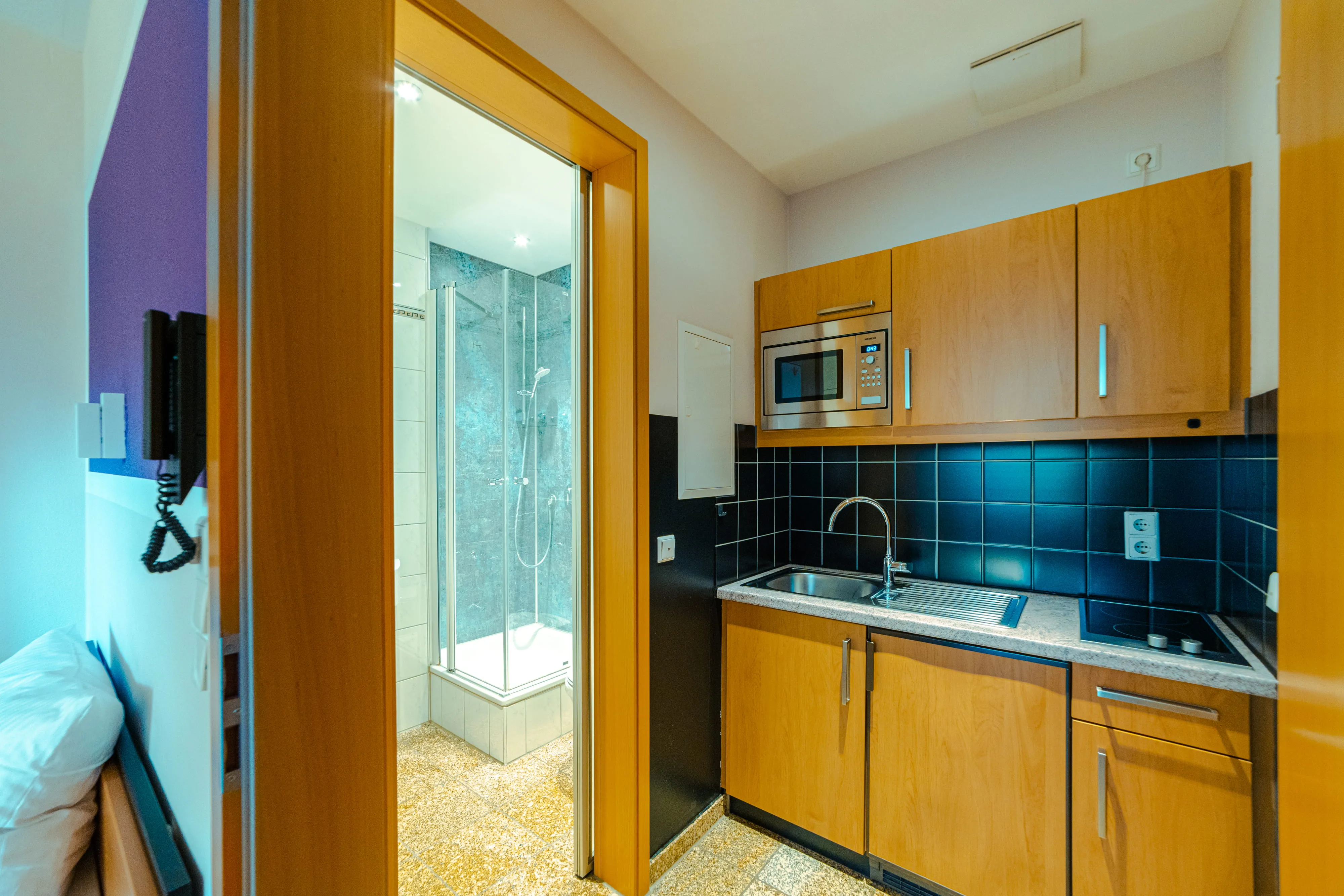 Compact kitchen area with wooden cabinets, a sink, a microwave, and a stovetop next to a doorway leading to a bathroom with a glass shower enclosure.