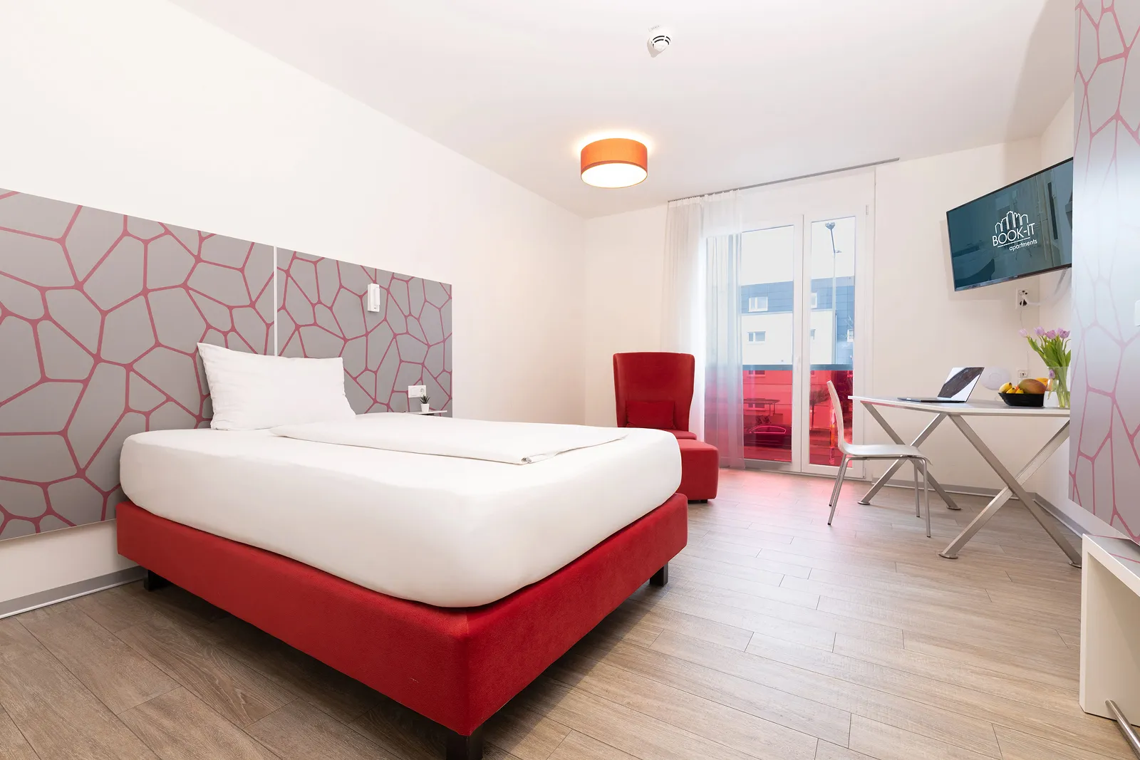 Modern hotel room with a red bed frame, white linens, red armchair, desk with laptop, and wall-mounted TV.