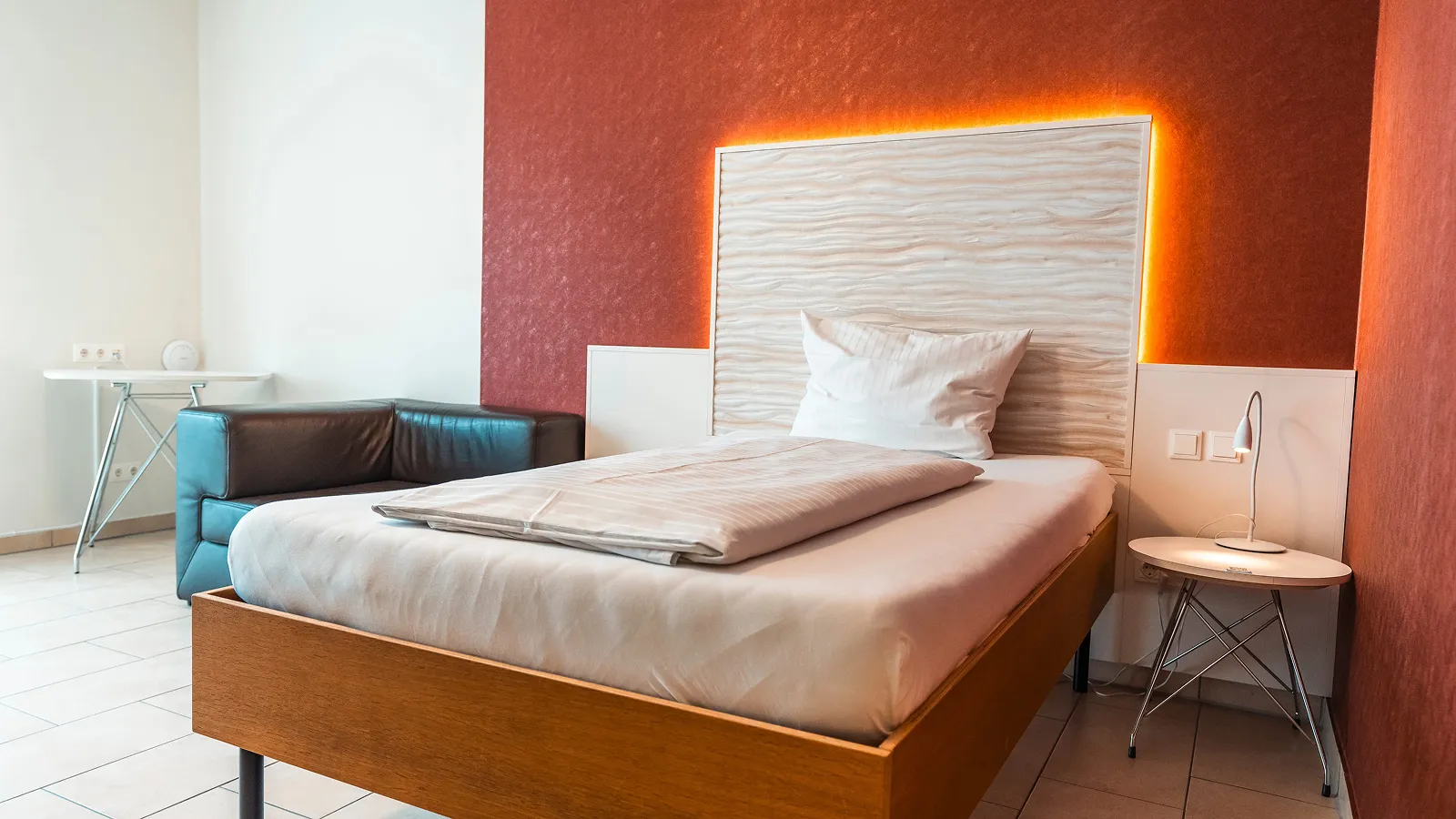 Modern single bed with white linens, a white textured headboard backlit with orange lighting, next to a small white table lamp and a black leather armchair in a tiled room.