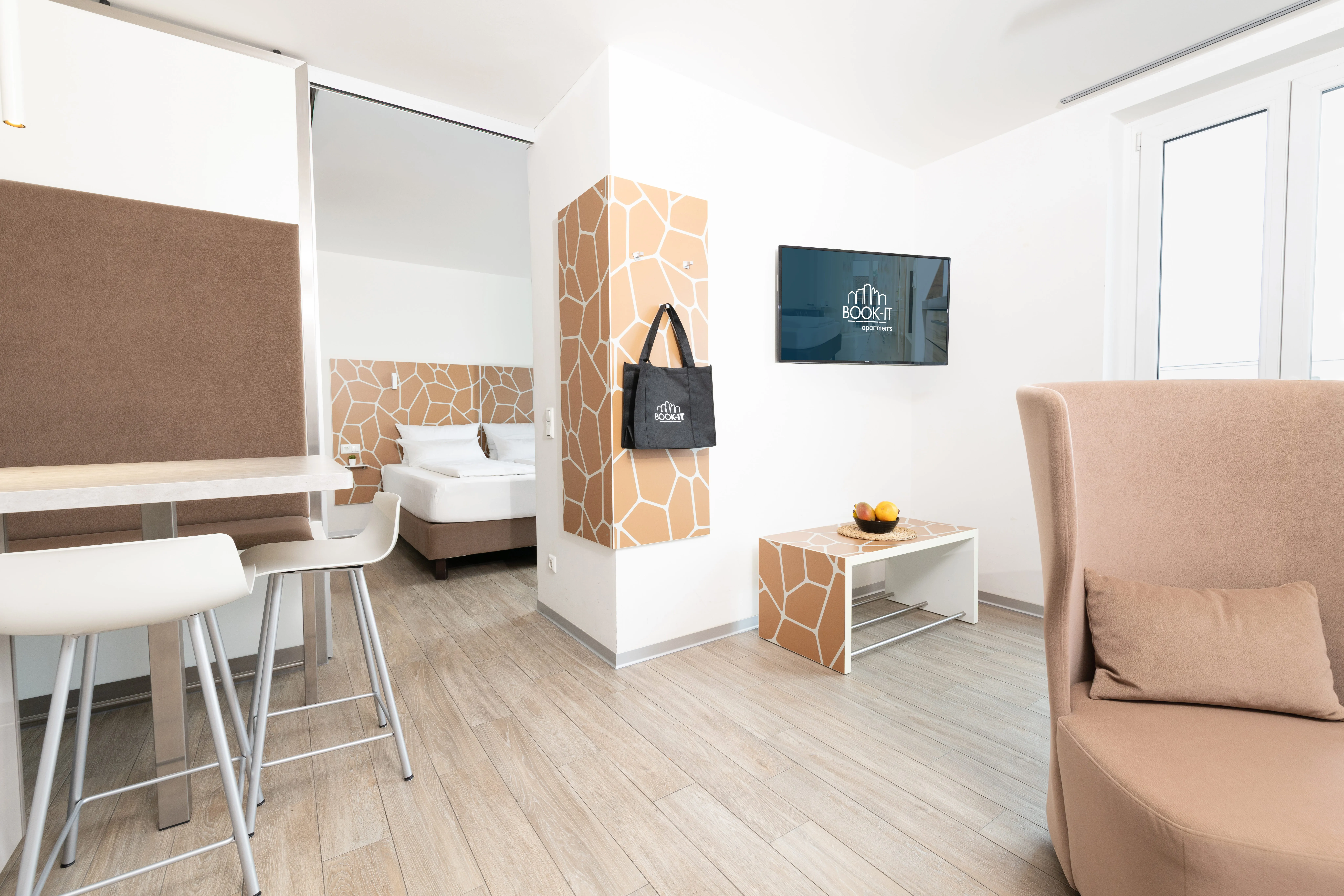 Bright modern apartment living area with beige armchair, wall-mounted TV, dining table and chairs, and a bedroom visible through a glass door.