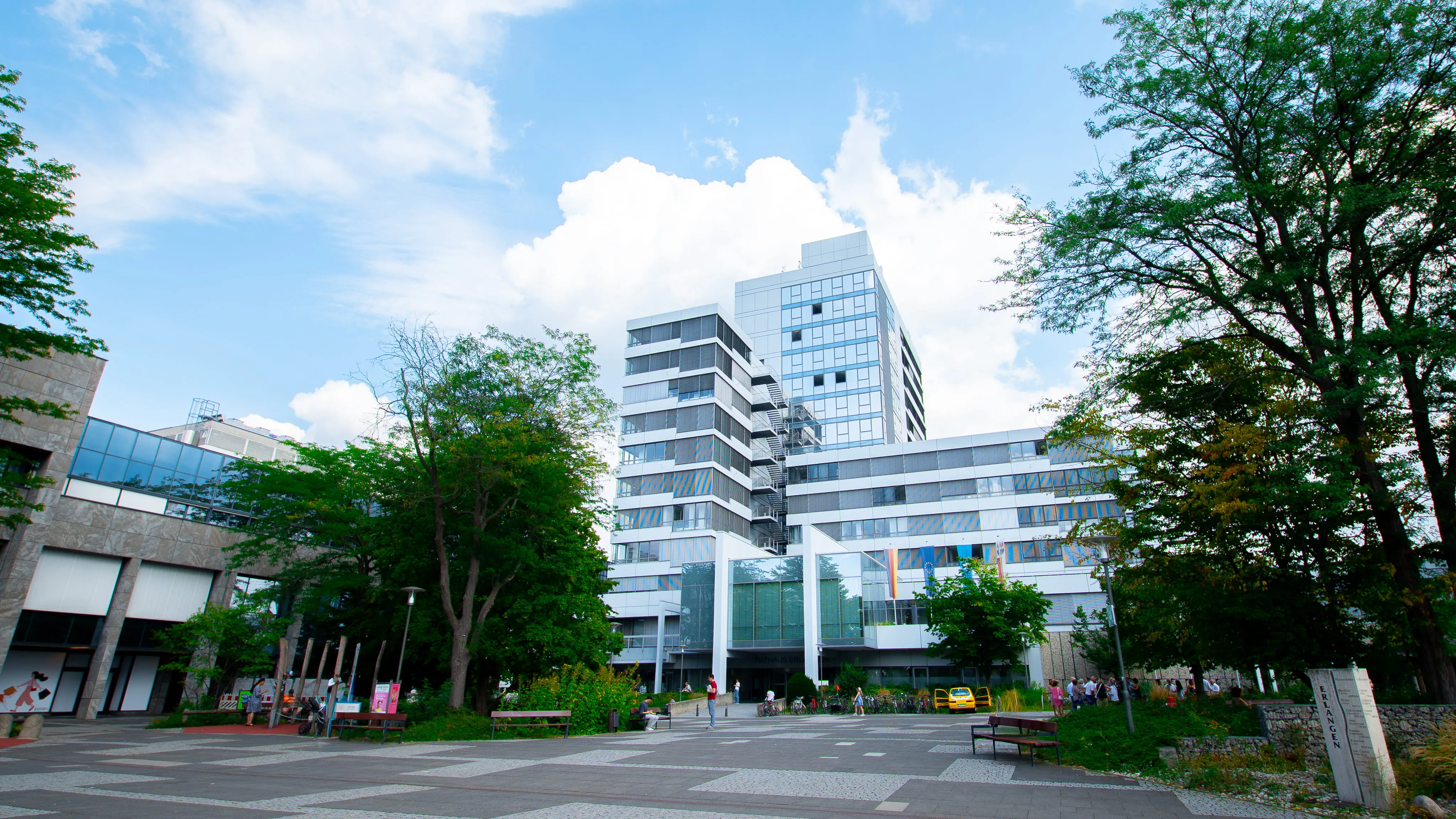 Modern multi-story white and glass building surrounded by trees under a blue sky with clouds.