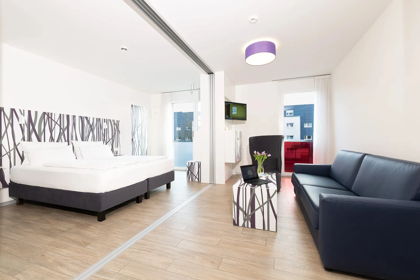 Modern hotel room with a double bed, a black sofa, a patterned coffee table with laptop and tulips, and a wall-mounted TV.