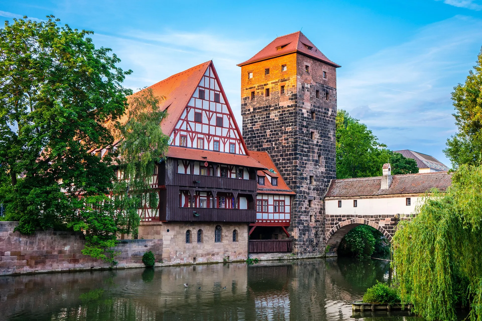 Historic half-timbered building and stone tower by a calm river with surrounding green trees under a blue sky.