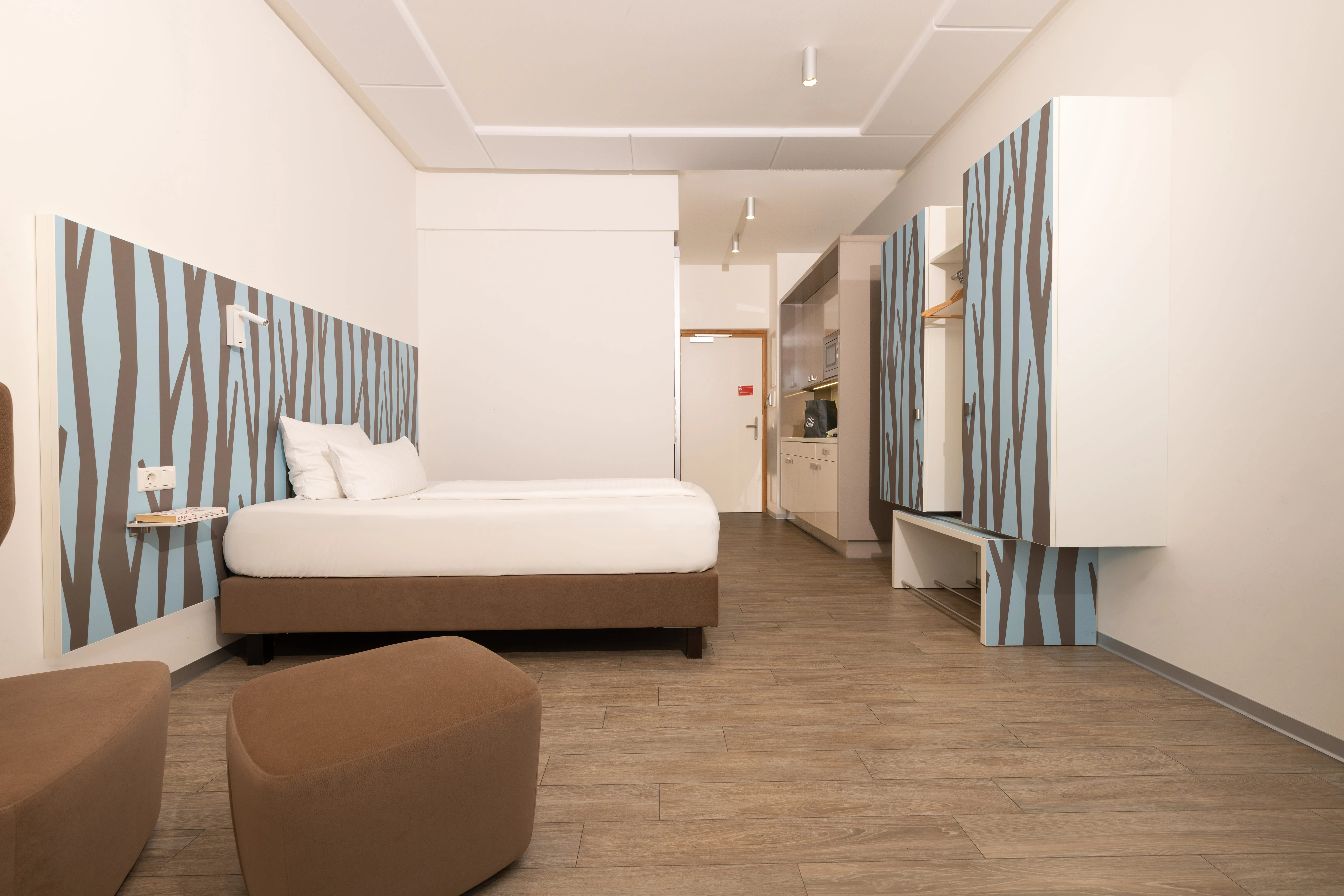 Modern hotel room with a double bed, brown ottomans, patterned headboard and wardrobe, and a kitchenette near the entrance.