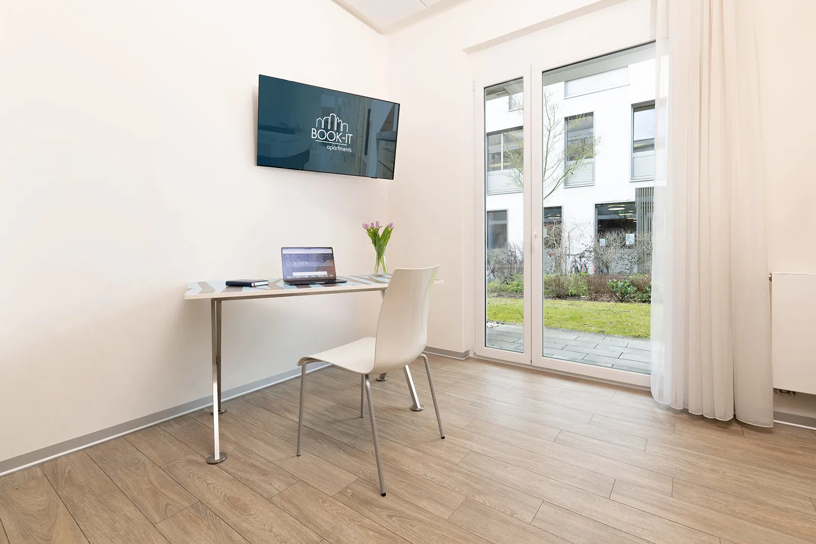 Minimalist workspace with a wall-mounted TV displaying 'BOOK-IT apartments', a table with a laptop and flowers, and glass doors overlooking a garden.