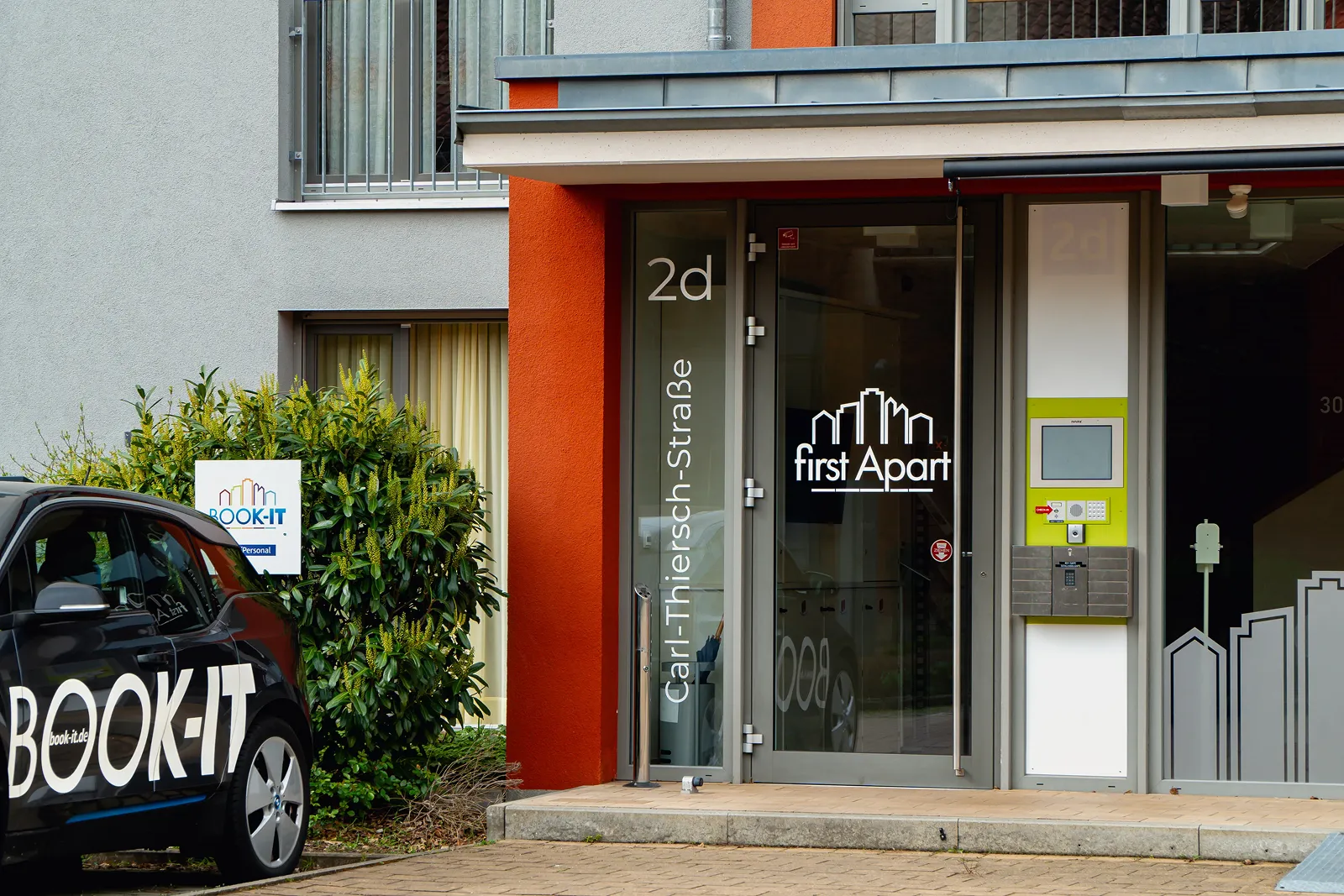 Entrance of a modern apartment building with 'first Apart' logo on glass door, located at Carl-Thiersch-Straße 2d, and a black car with 'BOOK-IT' branding parked nearby.