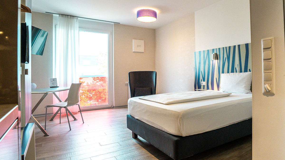 Modern hotel room with a large double bed, white bedding, patterned headboard and cabinets, wood-flooring, and a wall-mounted TV.