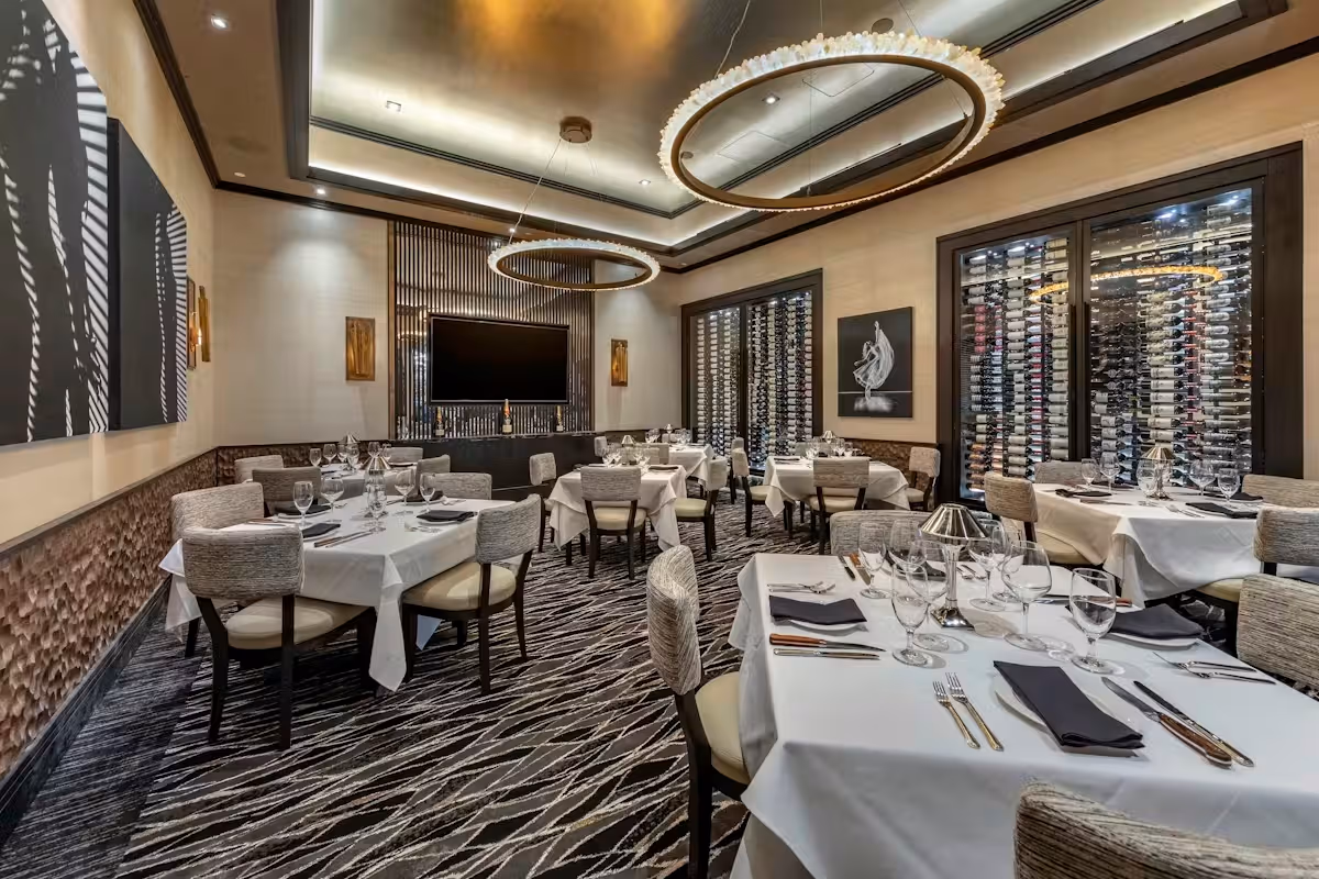 Elegant restaurant dining room with white tablecloths, modern chandeliers, and a wall of wine bottles.