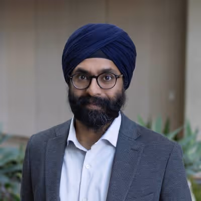 Man wearing a navy blue turban, glasses, white shirt, and grey blazer standing outdoors with plants and a building in the background.