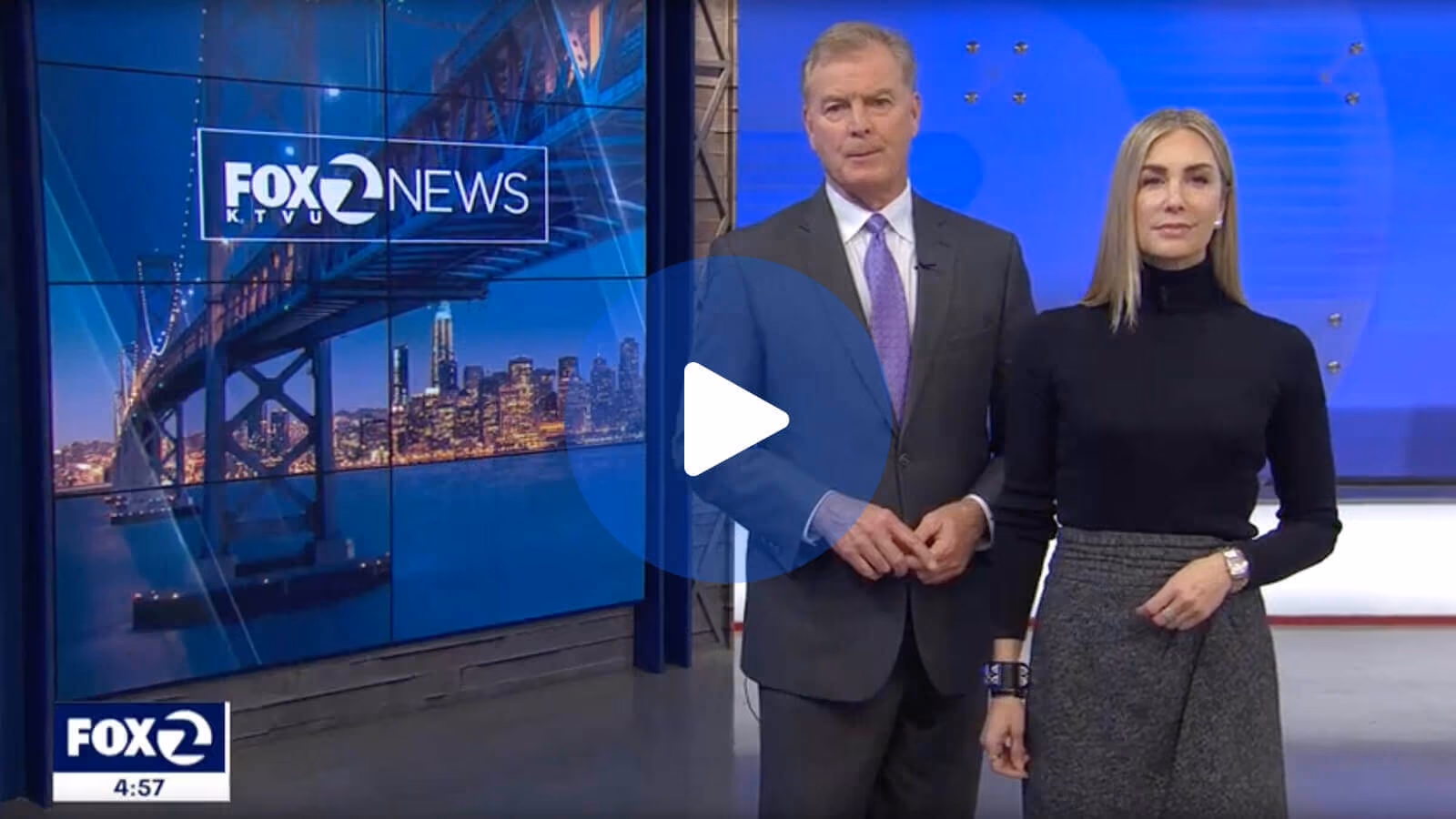 Two news anchors standing in a studio with a Fox 2 News screen showing a cityscape and bridge.
