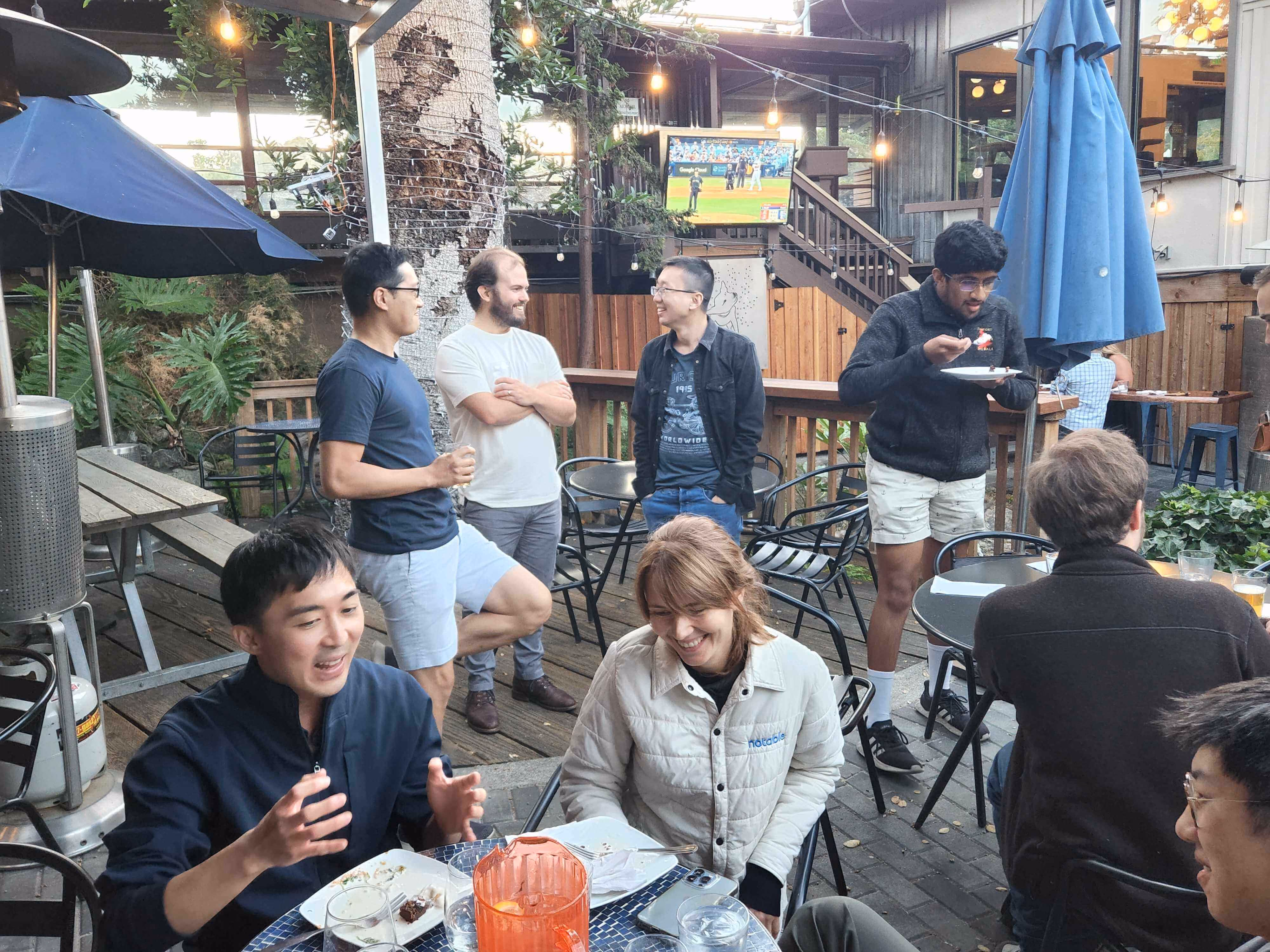 Group of people socializing and eating on a patio with a TV showing a baseball game.