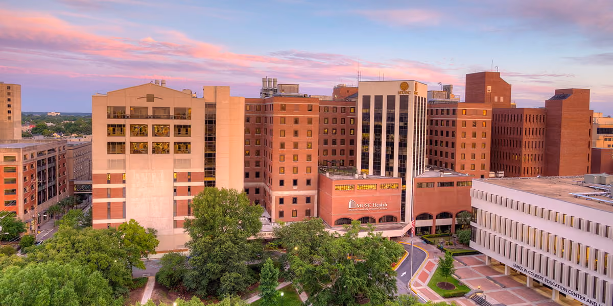 Wide view of MUSC Health hospital buildings with trees and a pink sky at sunset.
