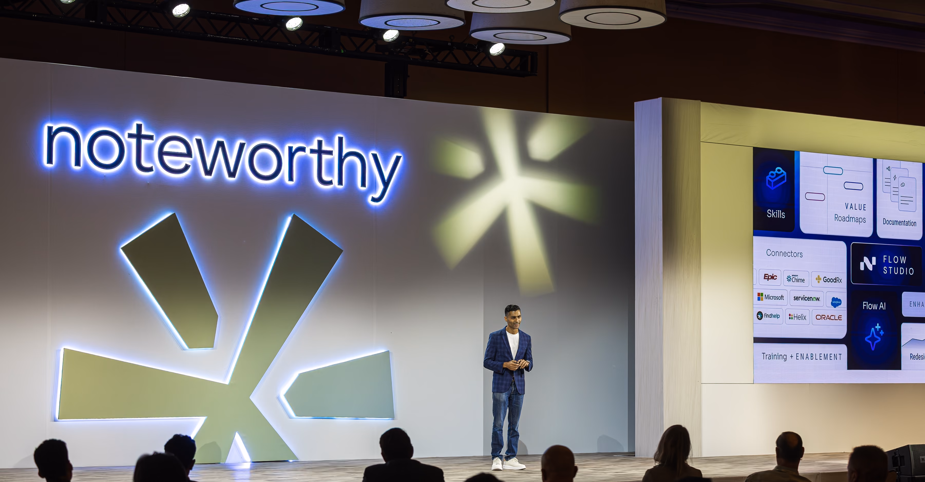 Speaker in casual outfit presenting on stage with large 'noteworthy' sign and screen showing flow studio features.